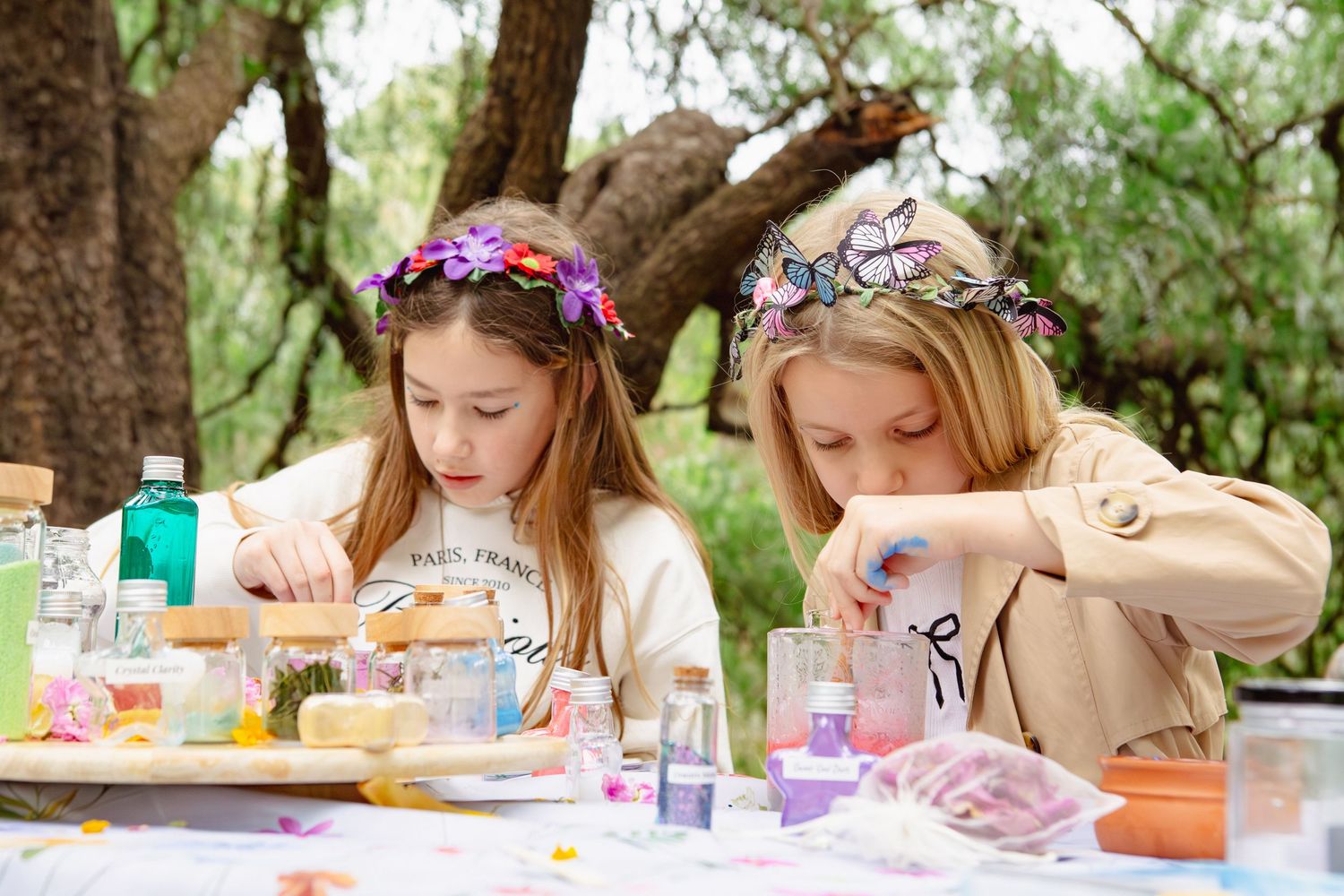 Two girls wearing flower crowns doing a craft activity at an outdoor table with colorful supplies.