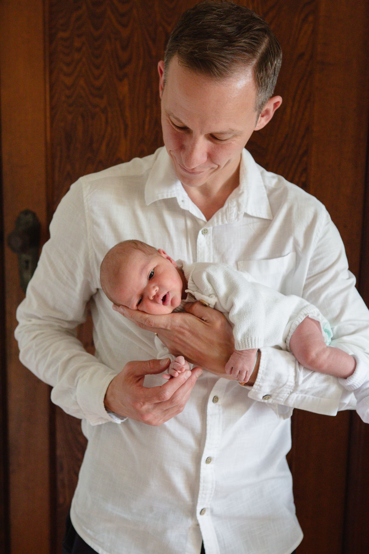 Smiling father in white shirt gently cradling his newborn baby in both hands, standing in front of a wooden door.