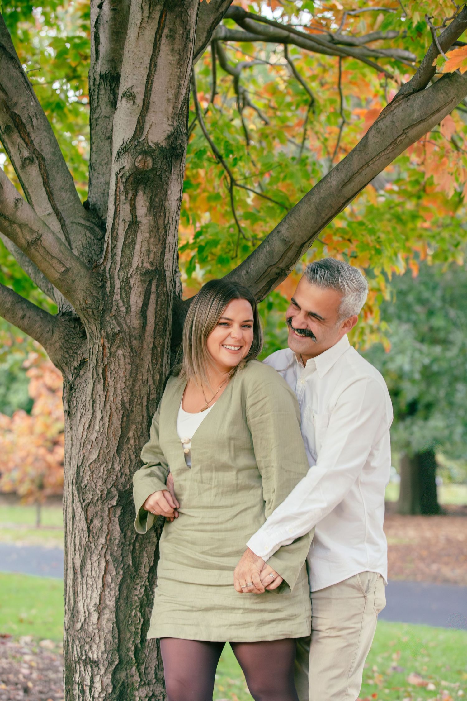 A smiling couple holds hands under a colorful autumn tree in a park, dressed in casual earth tones.
