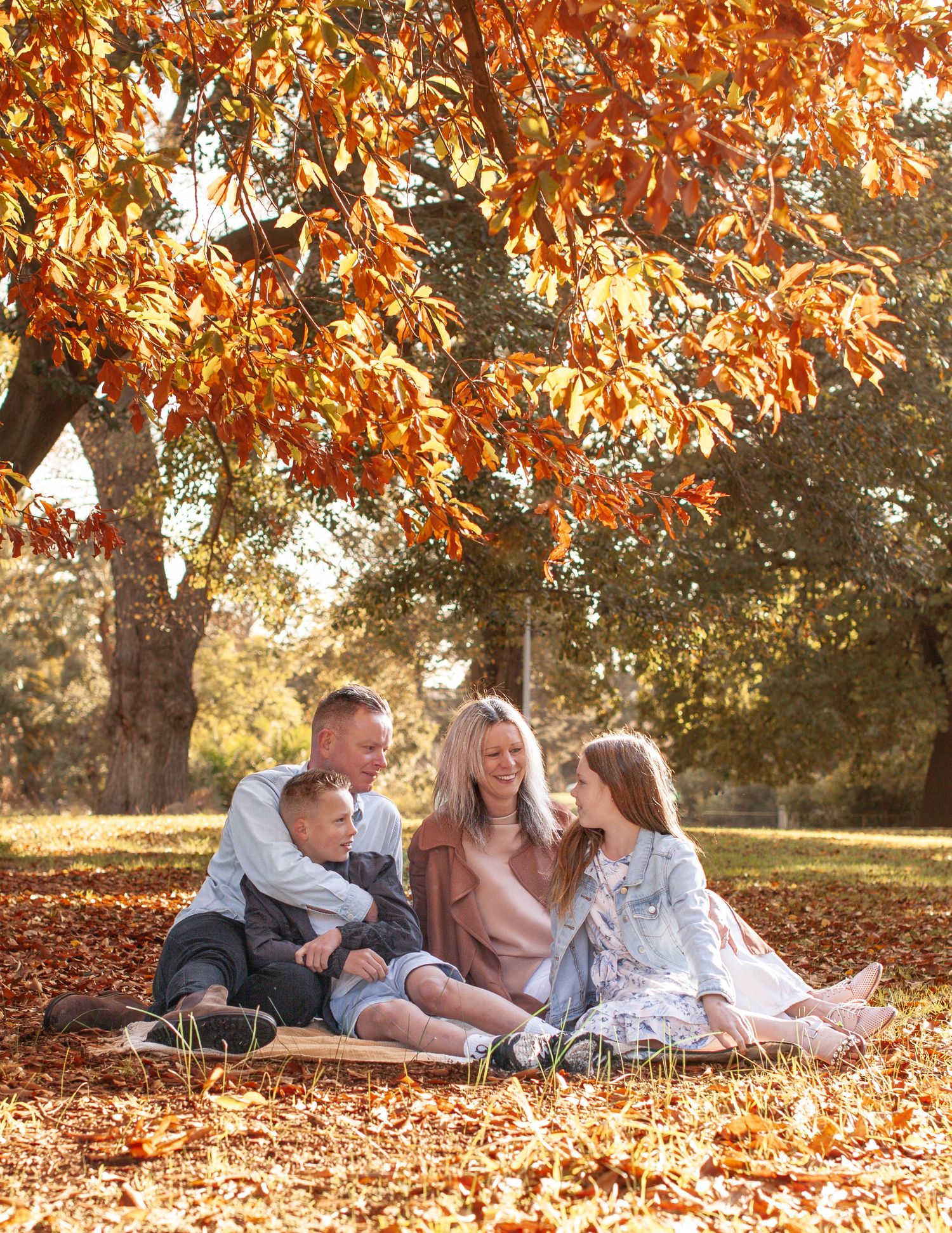 Family of four sitting together under vibrant autumn orange leaves in a sunlit park.