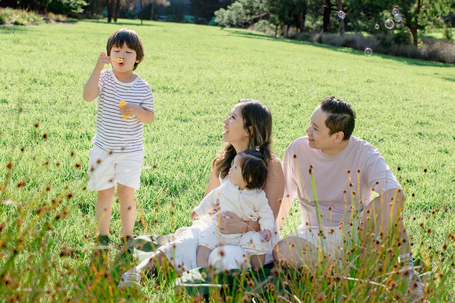 Family of four enjoying a sunny day in a green park, with a boy blowing bubbles while parents sit on grass.
