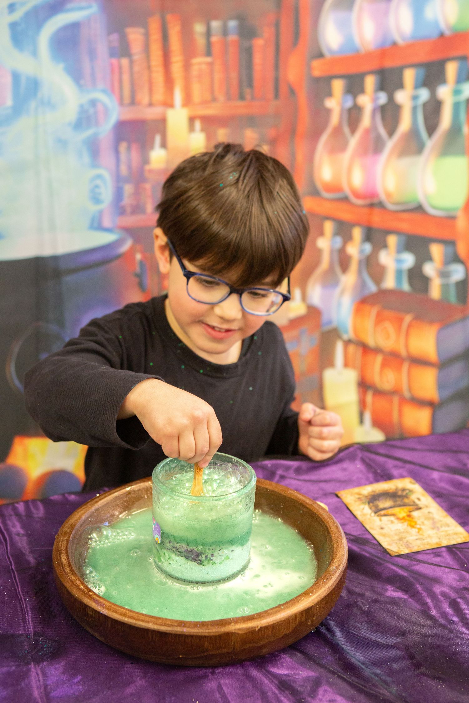 Young boy with glasses stirring green liquid in a glass jar at a magical potion-making activity station.