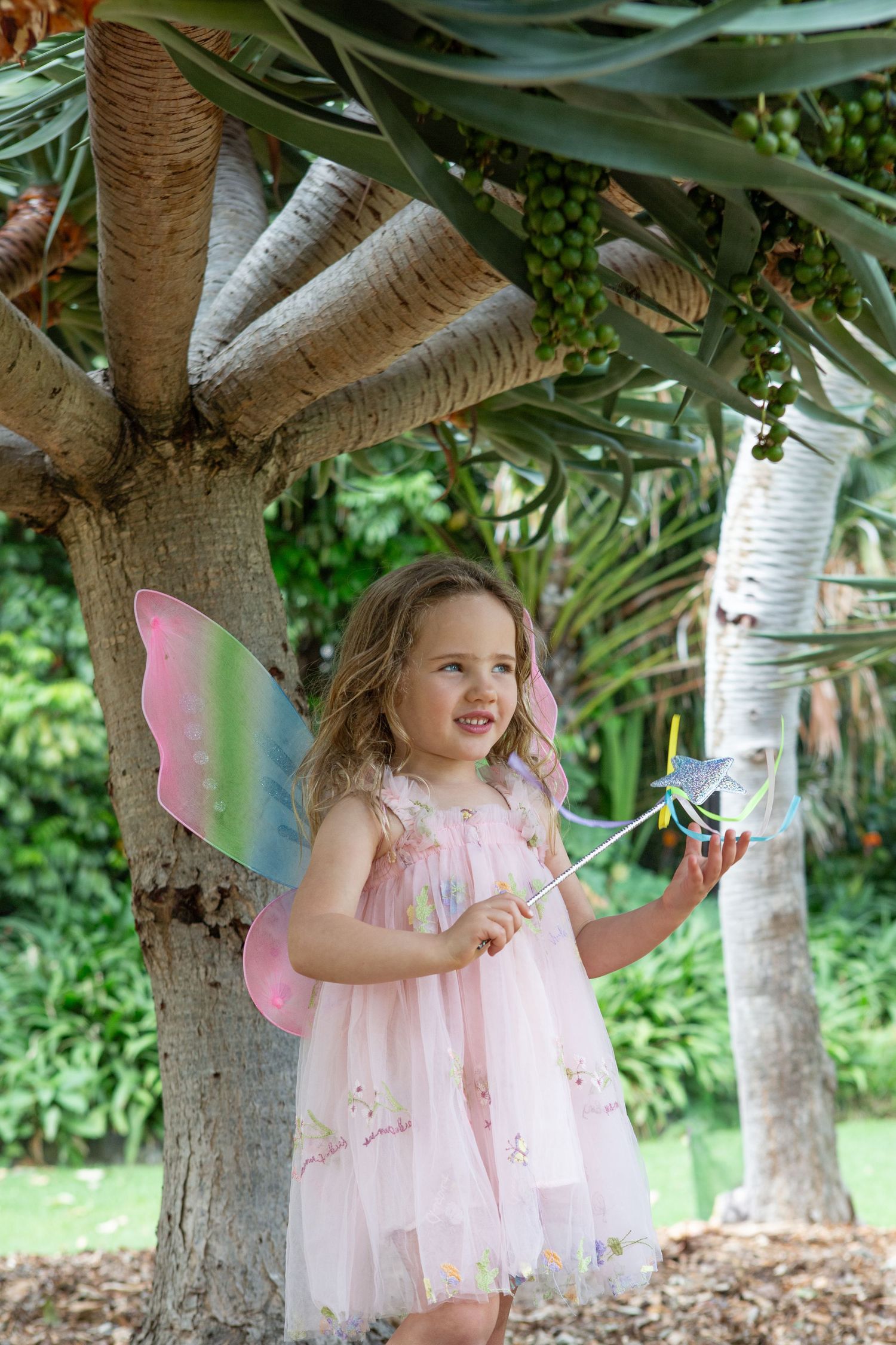 A young girl in a pink fairy dress with colorful butterfly wings holds a magic wand while standing under a tropical tree.