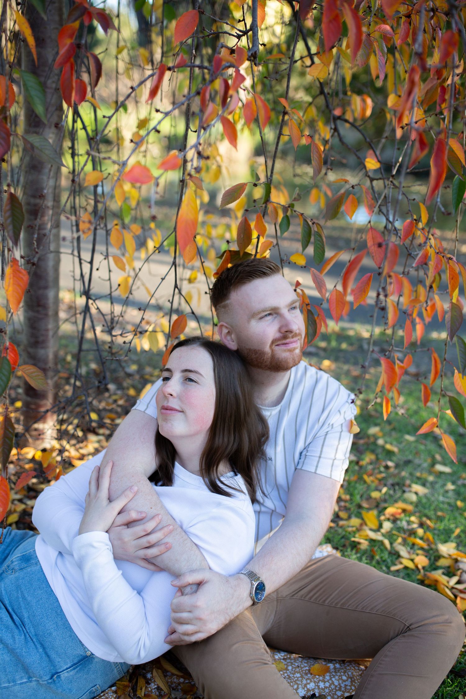 Couple sitting together under colorful autumn tree, man embracing woman from behind, surrounded by red and orange fall foliage.