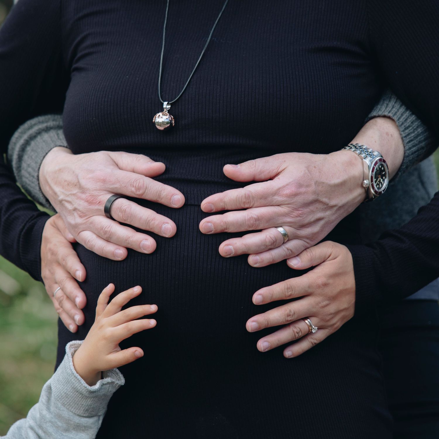 Pregnant woman in black dress with two adults and a child placing hands on her baby bump outdoors.
