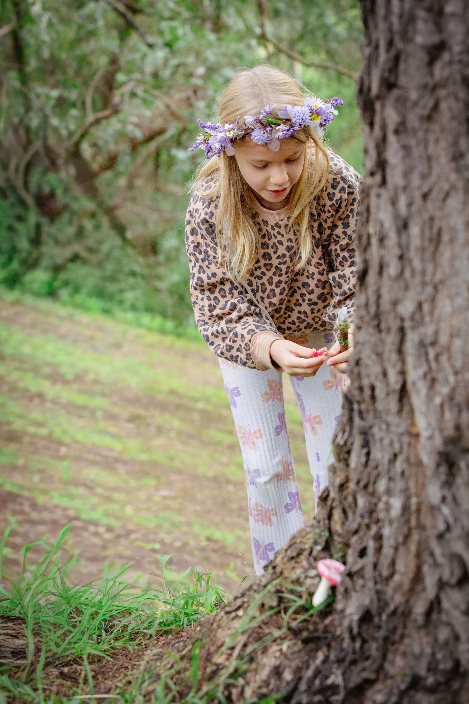 Blonde girl with flower crown discovering a tiny mushroom near a tree trunk in a forest.