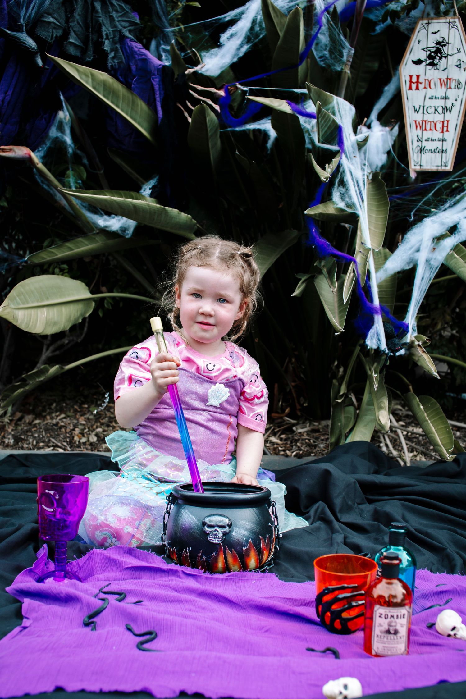 Young girl in pink dress stirs a Halloween witch cauldron with a wand surrounded by spooky decorations.