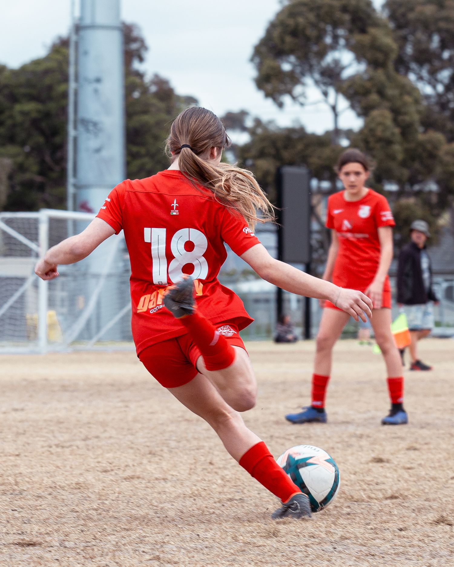 Female soccer player #18 in red uniform kicking ball during outdoor match with teammate in background.