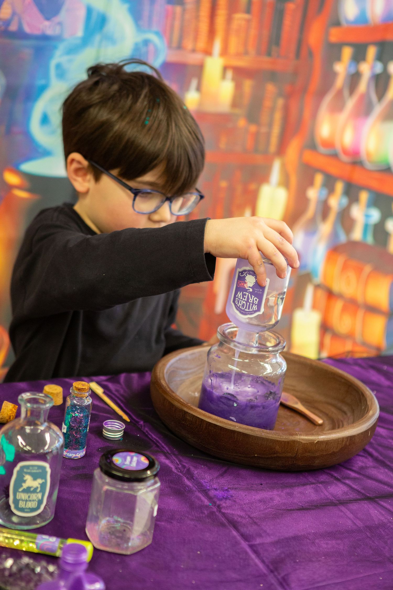 Young boy with glasses pouring purple potion into jar at a magical wizard-themed activity table.