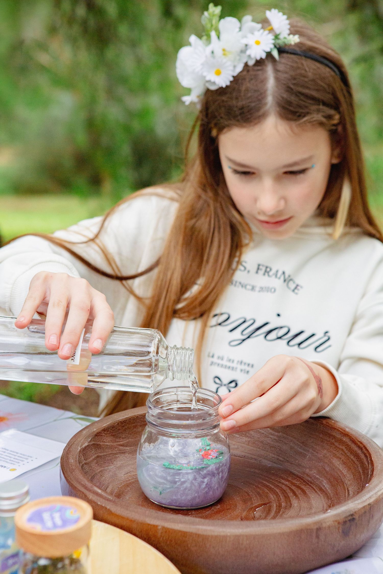 Girl with flower crown pouring water into a jar outdoors, crafting a nature project at a wooden table.