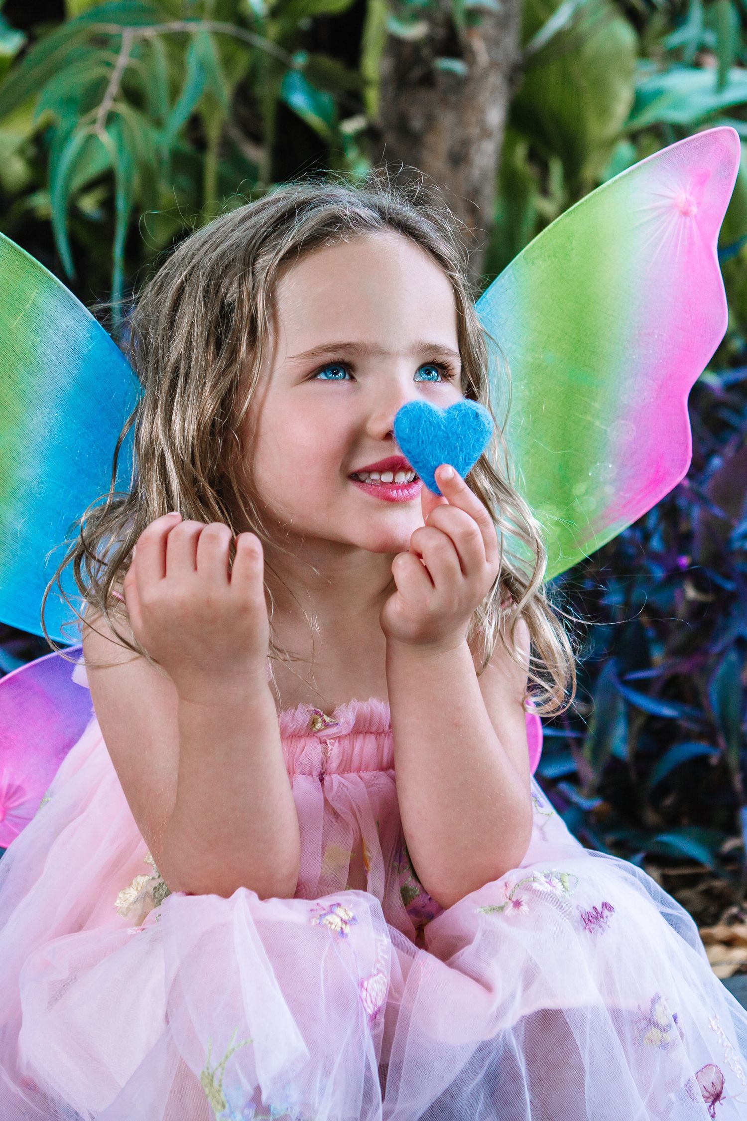 Young girl in pink fairy costume with rainbow wings holding a blue heart, smiling outdoors.