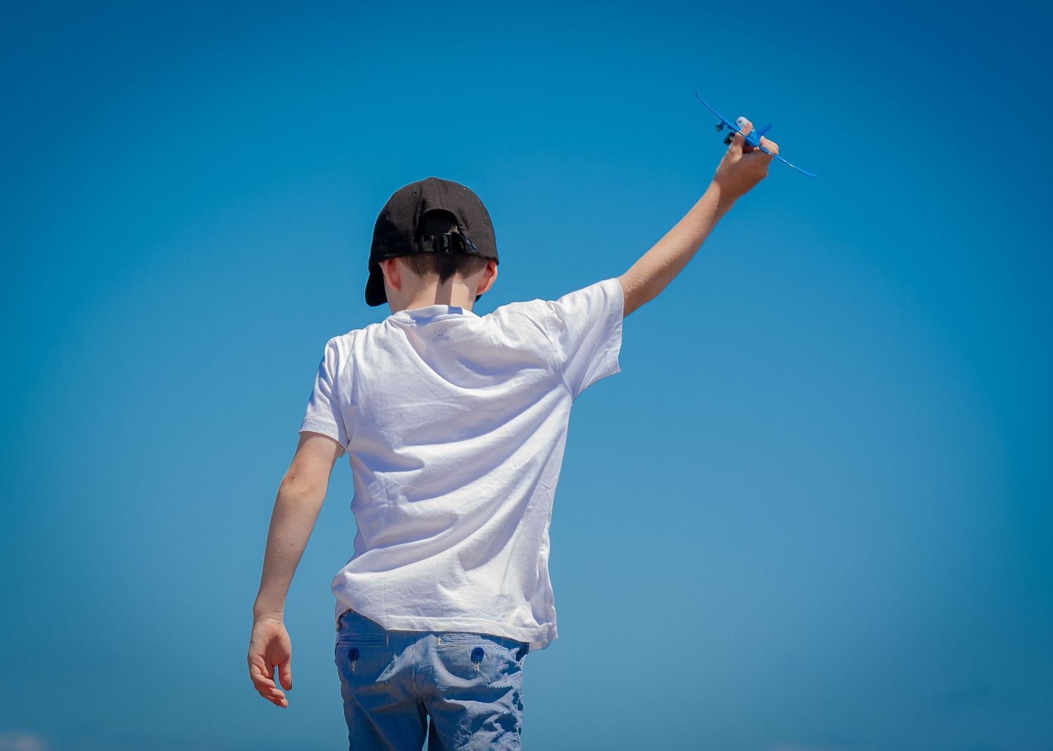 Child in white t-shirt holding a toy airplane up against a clear blue sky.