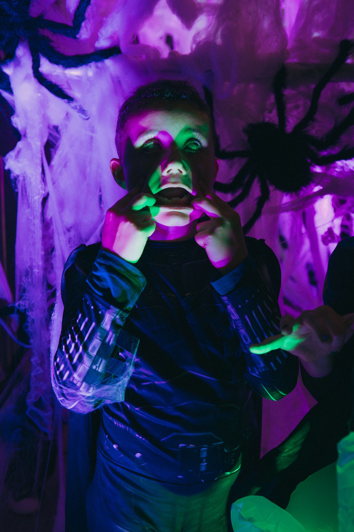 Boy in Halloween costume poses with green face paint under purple neon lights with spider decorations.