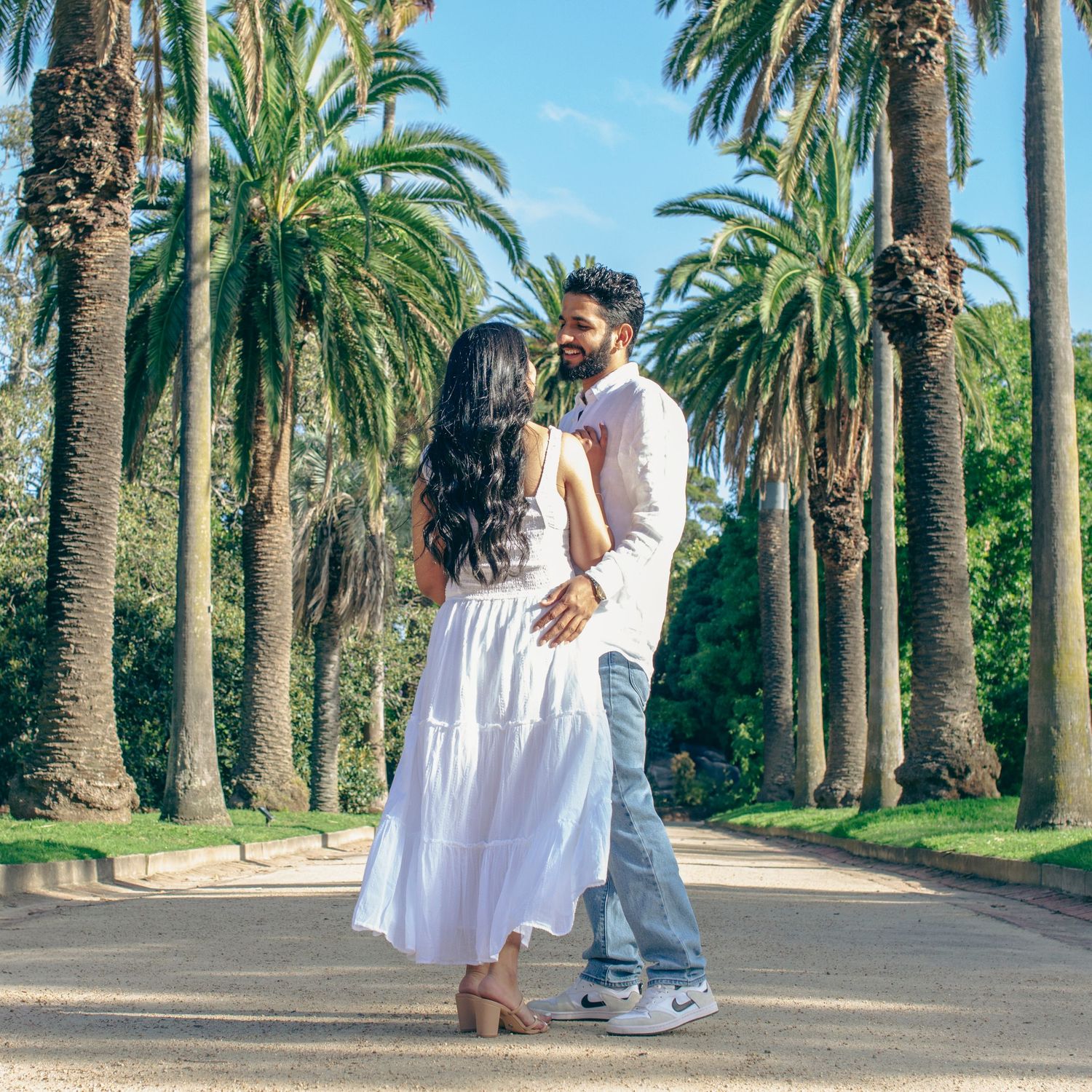Couple dancing on a palm tree-lined path in a sunny park, woman in white dress, man in casual white shirt and jeans.