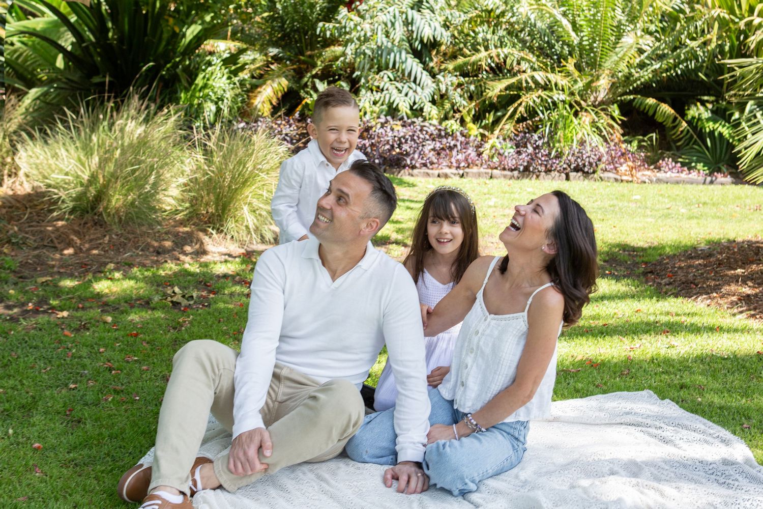 Happy family of four enjoying a sunny picnic on green grass in a lush garden park setting.