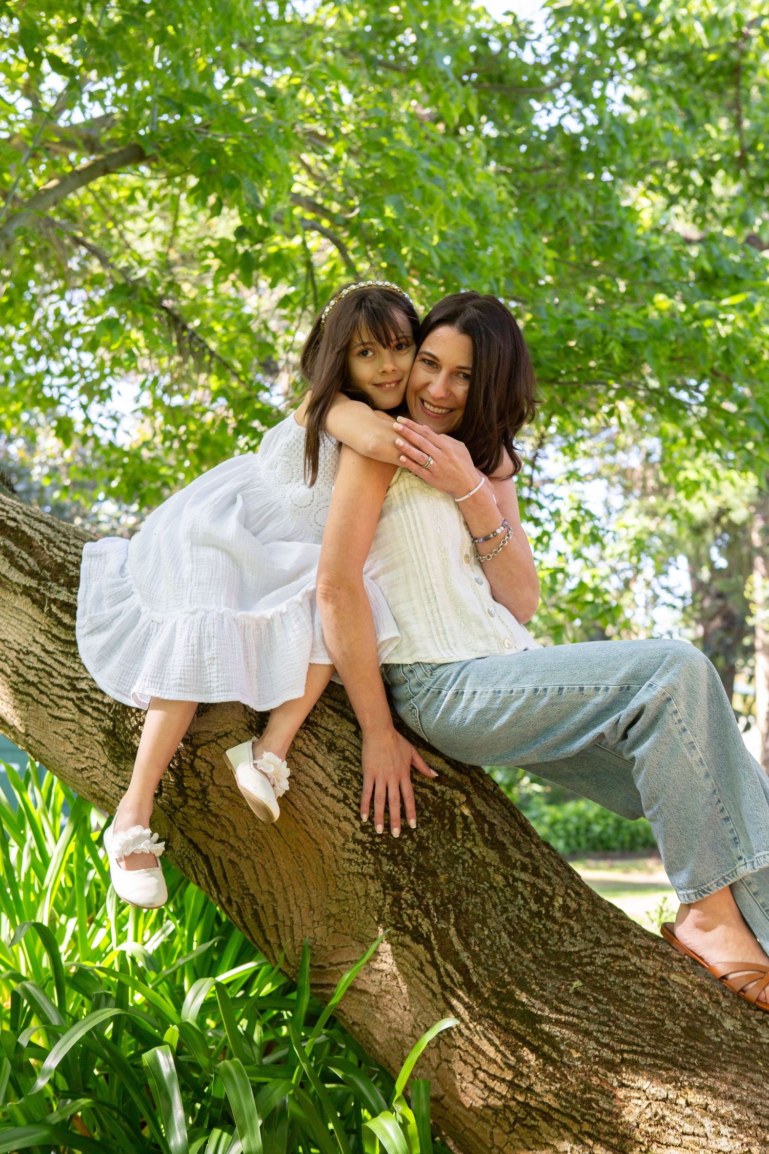 A mother and young daughter share a warm embrace while sitting on a large tree branch in a lush green park.