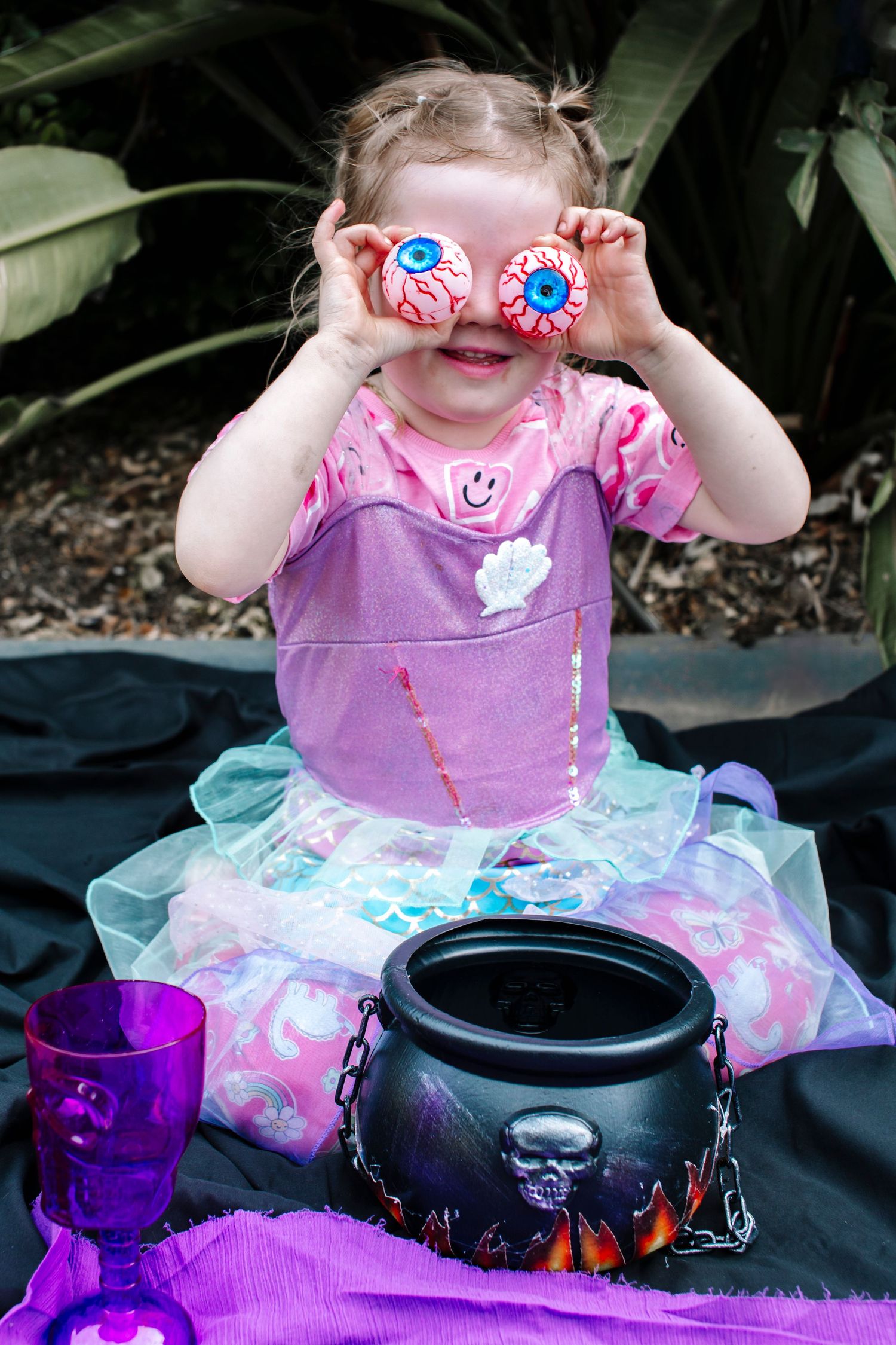 Toddler girl in fairy costume holds Halloween eyeball decorations over her eyes near a witch cauldron.