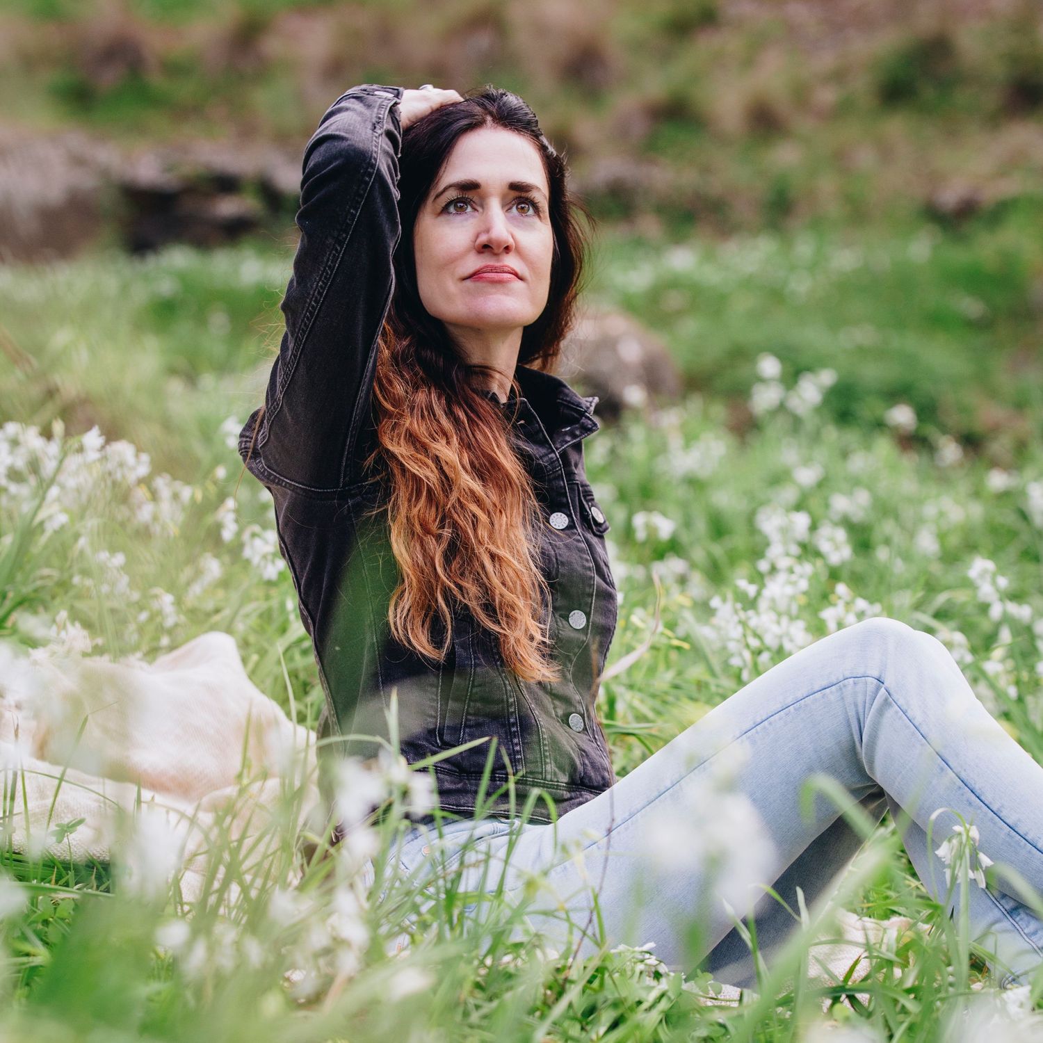 Woman with long hair sitting in a green meadow with white wildflowers, looking up thoughtfully on a cloudy day.