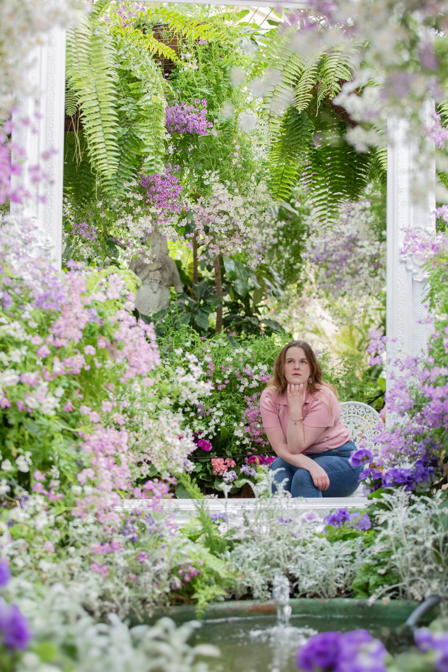 Woman sitting pensively in a lush garden gazebo surrounded by purple and pink blooming flowers and ferns.