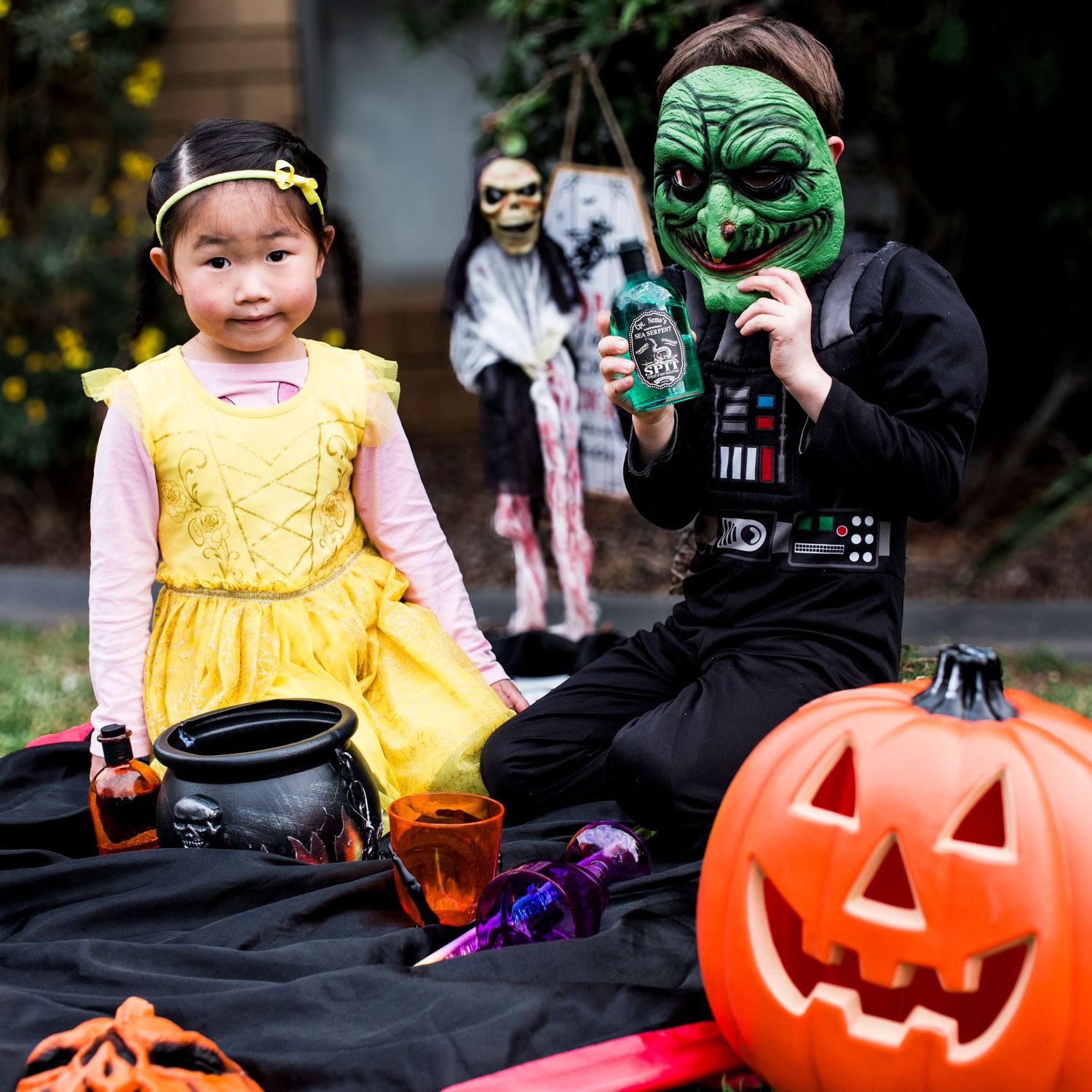 Two children in Halloween costumes sit outdoors with jack-o-lantern and cauldron decorations.
