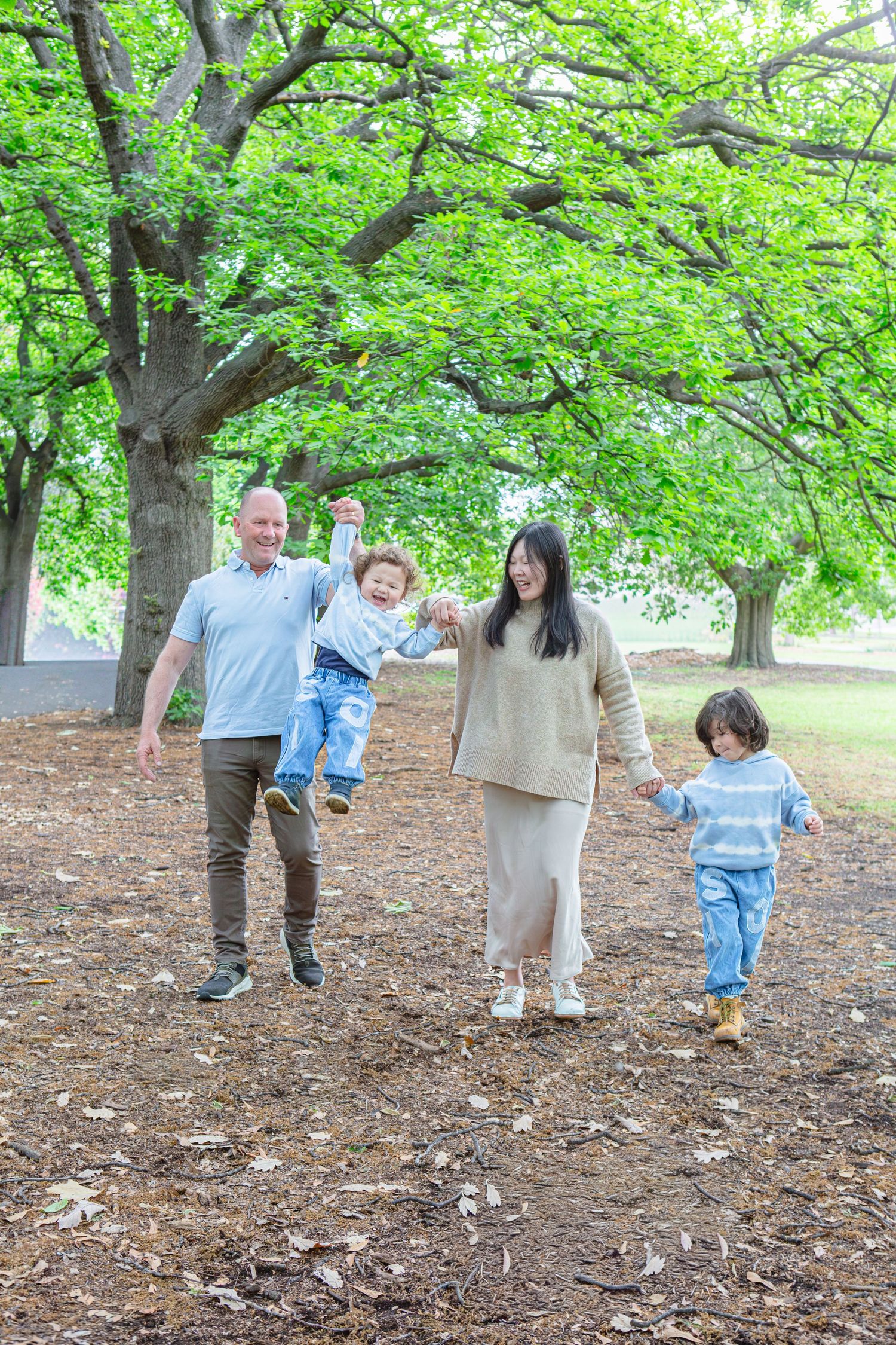 Family of four enjoying a walk in a lush green park, parents swinging toddler while older child holds hands.