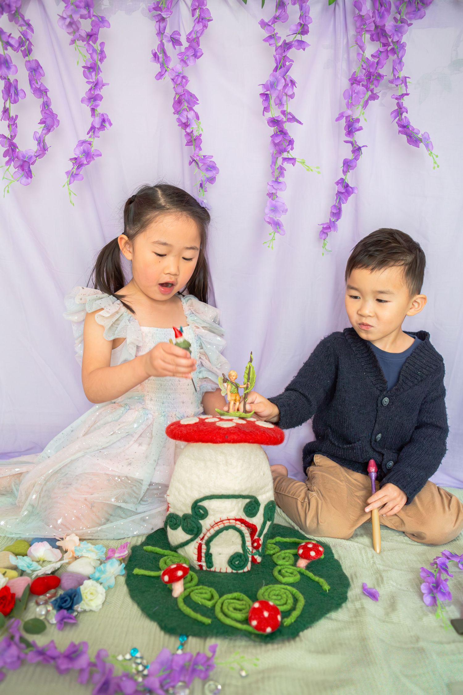Two Asian children play with a fairy mushroom house prop amid purple wisteria backdrop in a whimsical photo session.