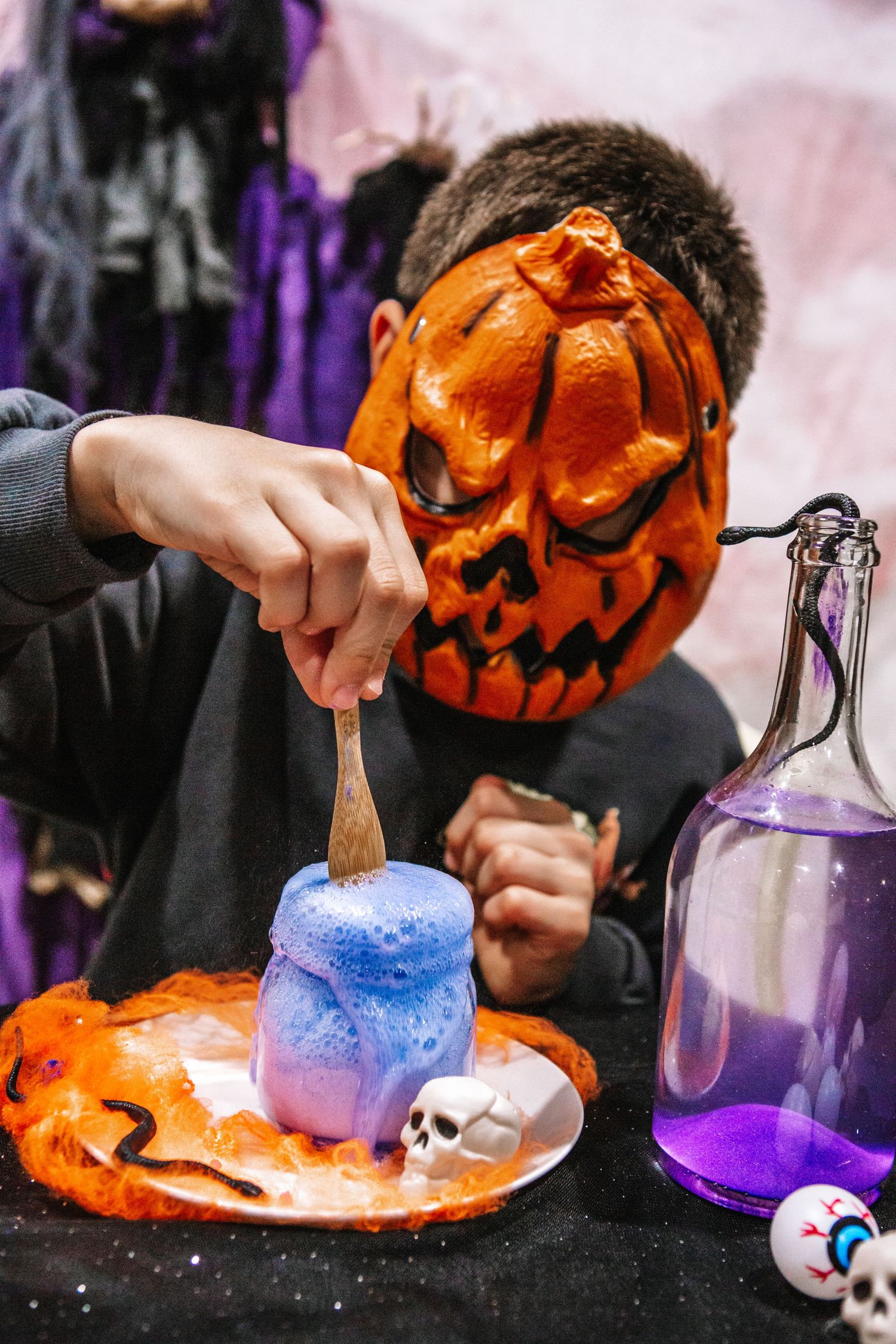 A child wearing a jack-o-lantern mask stirs a colorful Halloween potion at a decorated table.