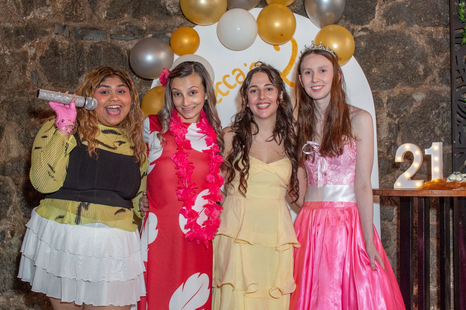 Four young women in colorful formal dresses pose together at a 21st birthday party with gold balloons.