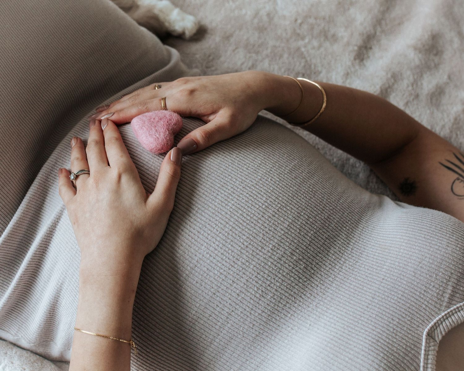 Two hands holding a pink heart-shaped crystal on a pregnant belly, with gold jewelry and a tattoo visible.