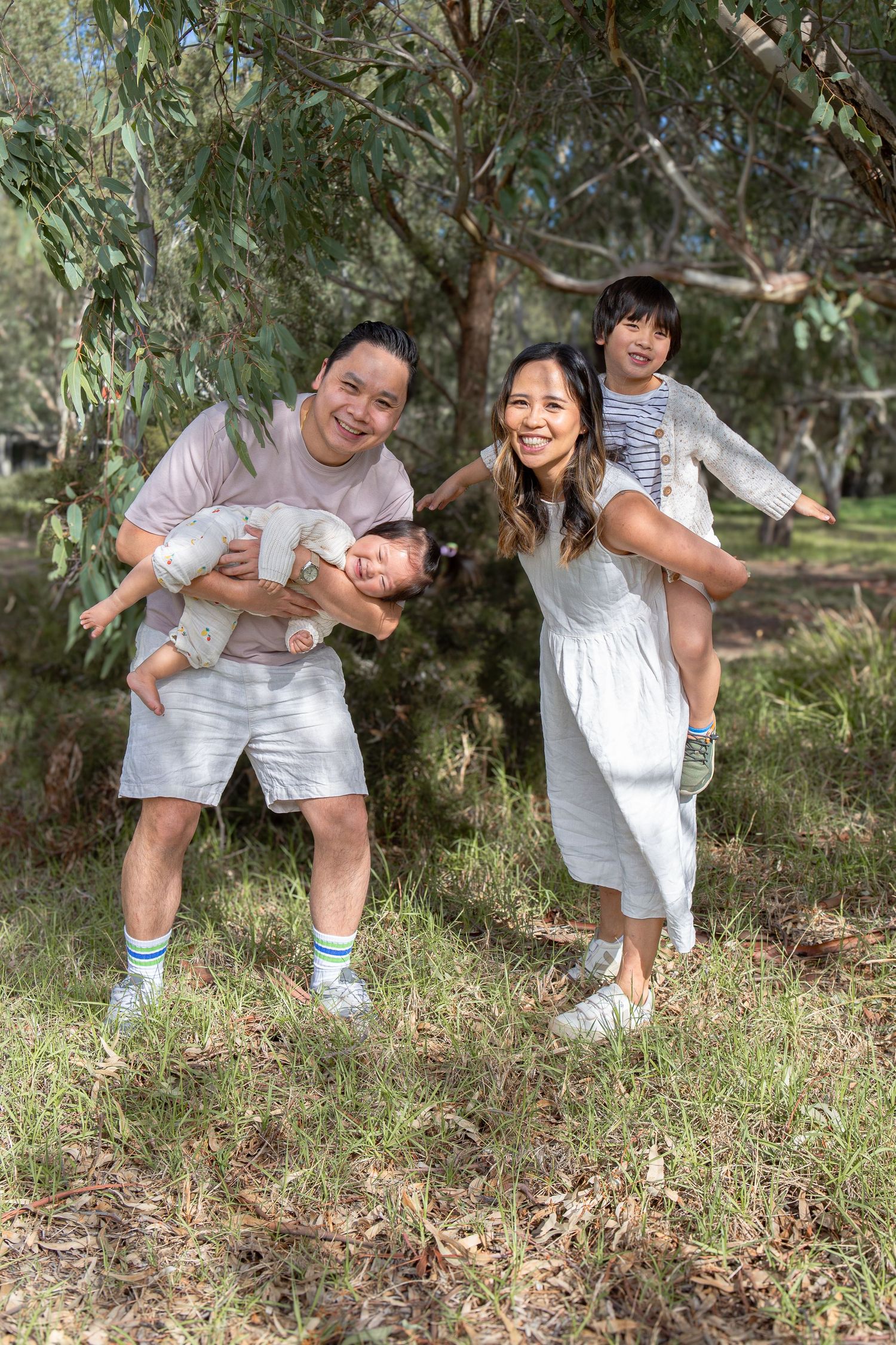 Happy Asian family with two children playing outdoors in a park, parents holding kids playfully among green trees.