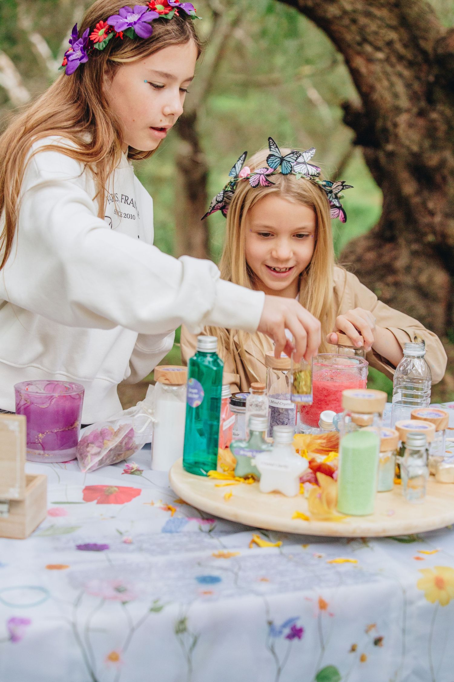 Two girls wearing flower crowns enjoy a colorful outdoor potion-making activity at a floral tablecloth-covered table.