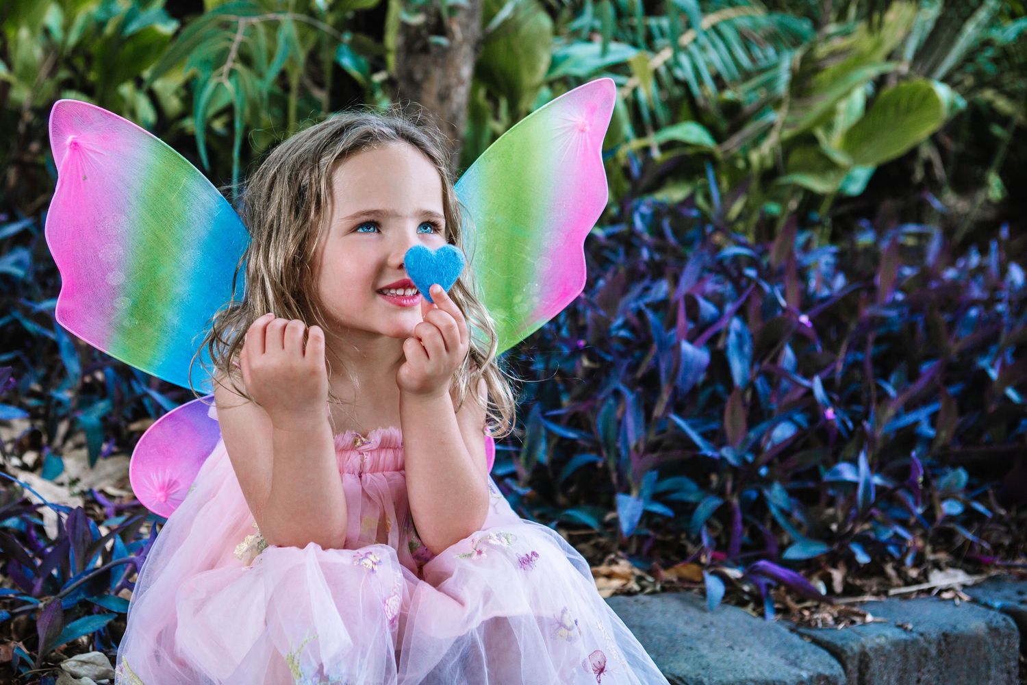 Adorable little girl in pink fairy costume with colorful butterfly wings holds blue flower outdoors.