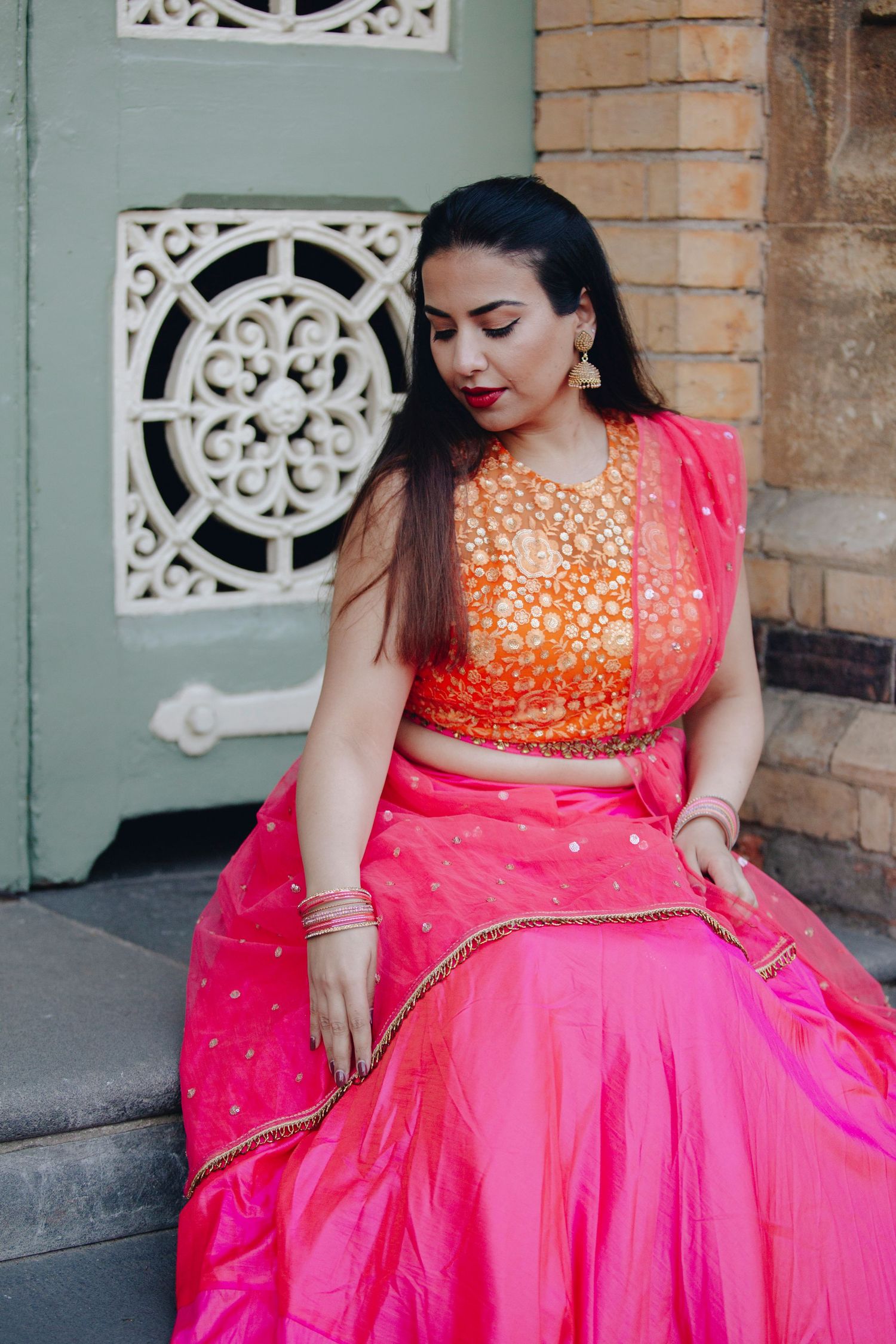 Woman in vibrant pink and orange Indian lehenga choli with gold jewelry, sitting gracefully near ornate architecture.