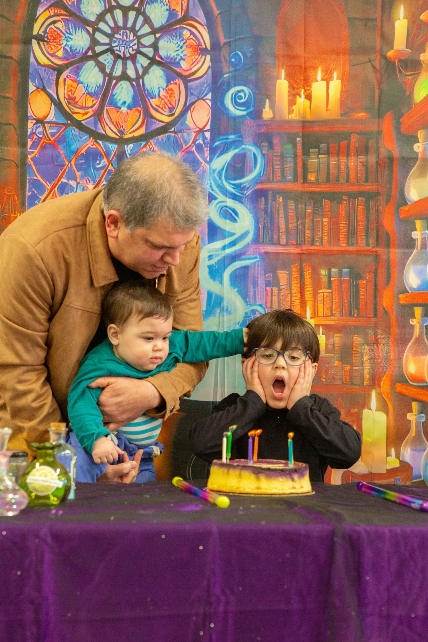 Child with glasses reacts with awe to birthday cake candles while man holds baby before colorful wizard library backdrop.