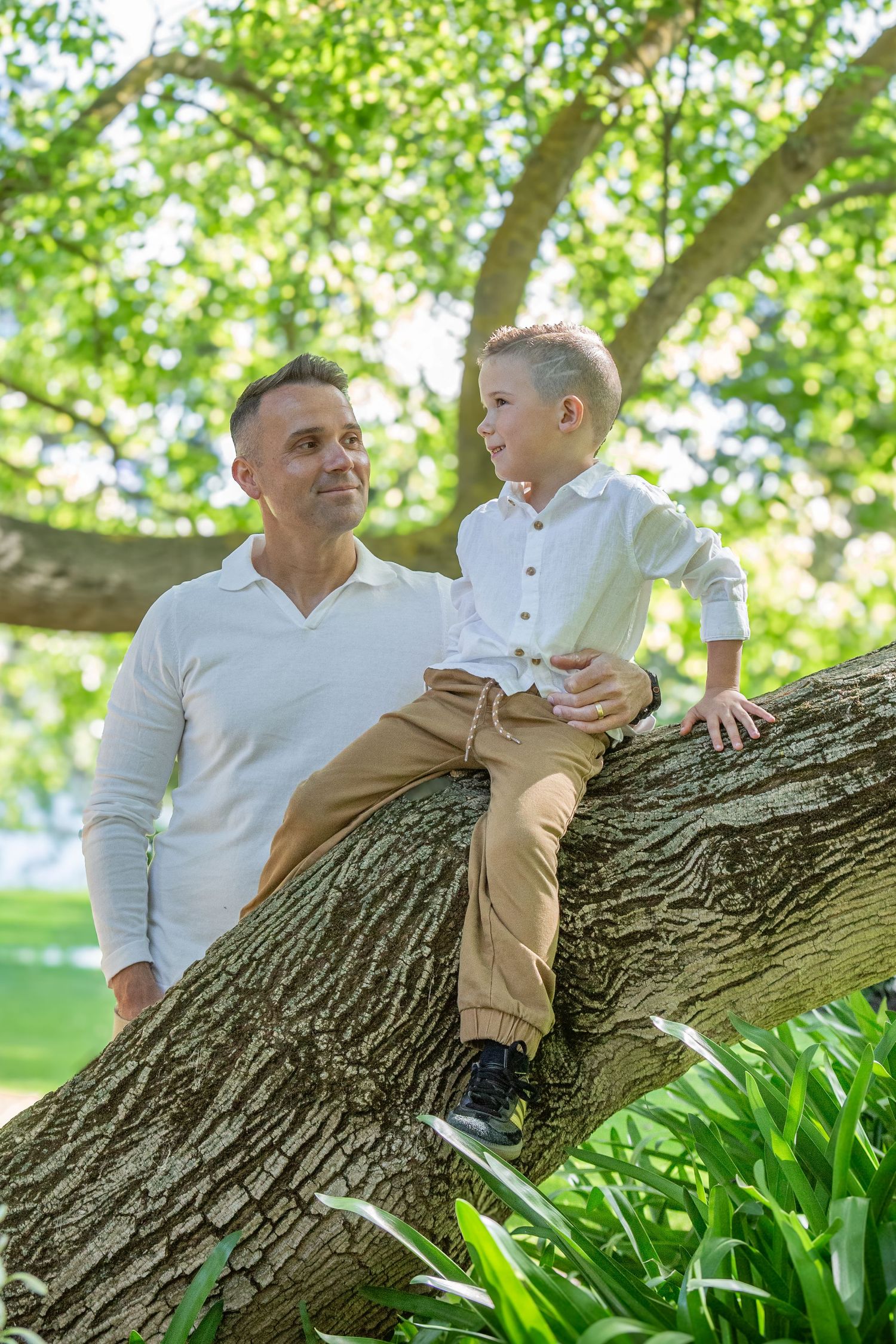 A father and young son share a joyful moment sitting on a large tree branch in a lush green park.