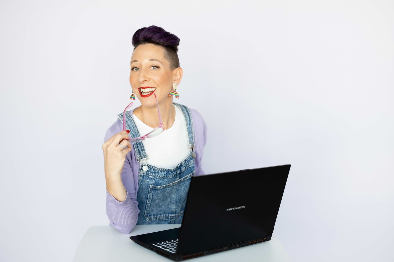 Creative woman with purple pixie cut wearing denim overalls, sitting at desk with laptop, holding pen and smiling on white background.