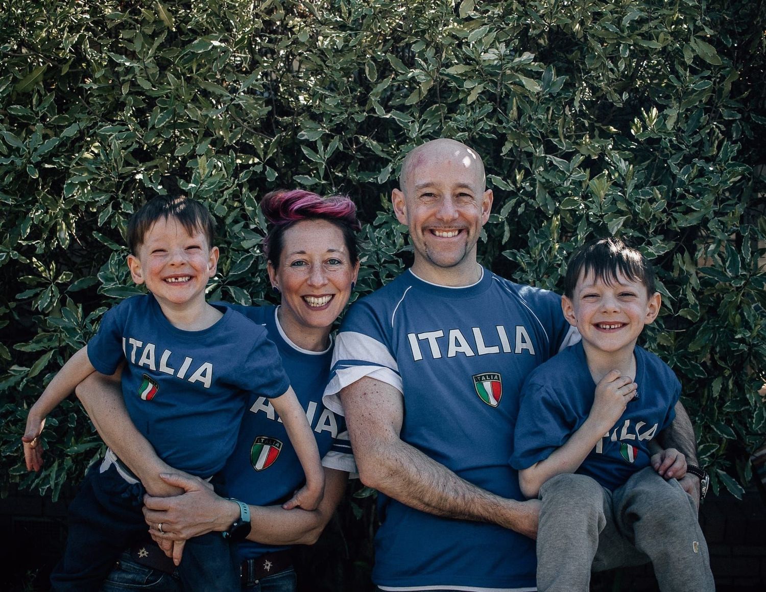 Happy family of four wearing matching Italia shirts posing in front of green foliage.