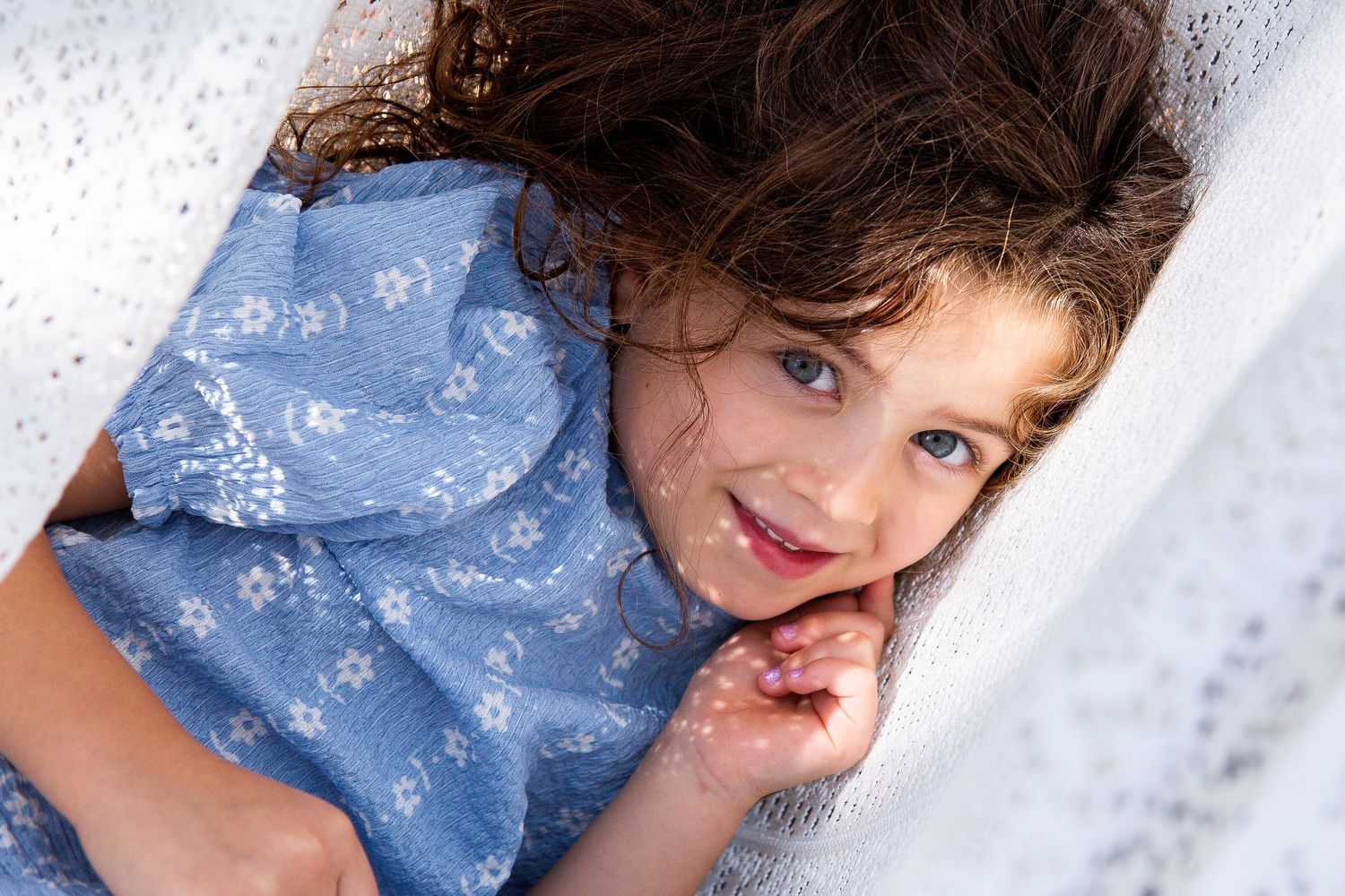 Curly-haired young girl in a blue floral dress lying in a white hammock smiling.