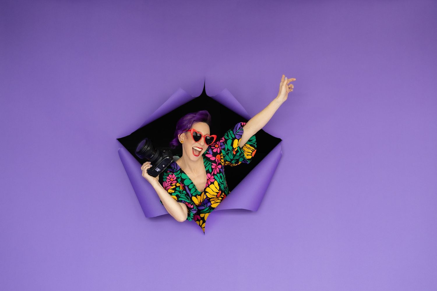 Joyful woman with purple hair in floral outfit peers through torn purple paper background.