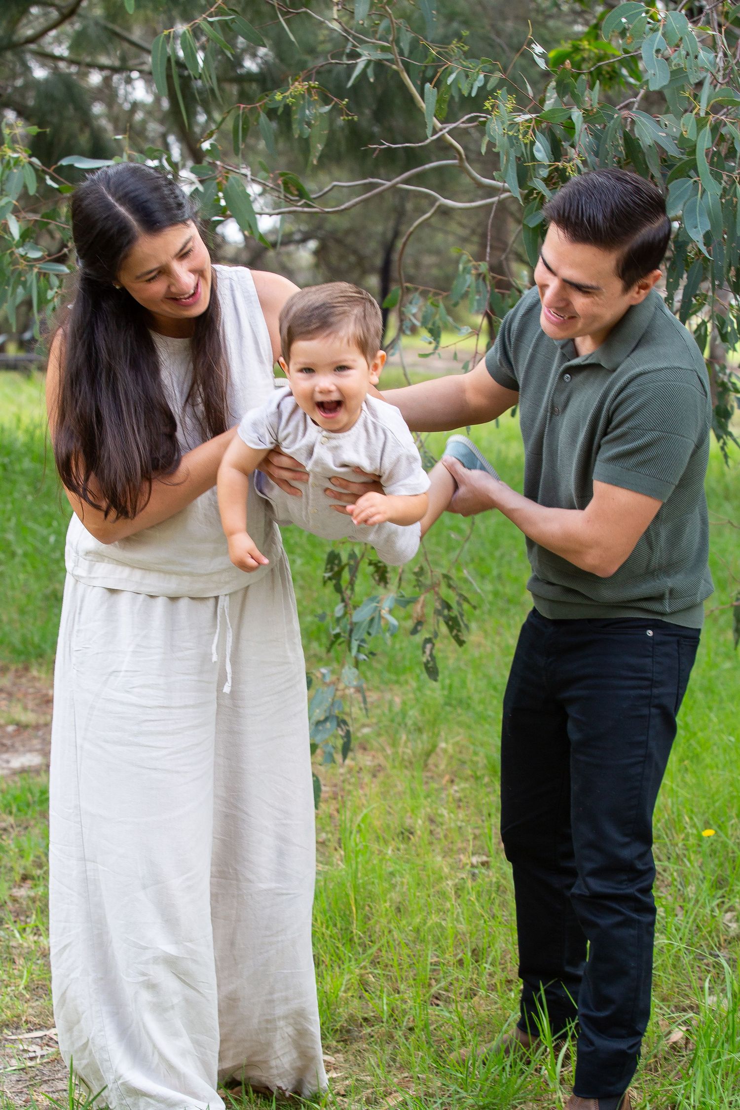 Happy young family with laughing baby enjoys outdoor portrait session under eucalyptus tree in park.