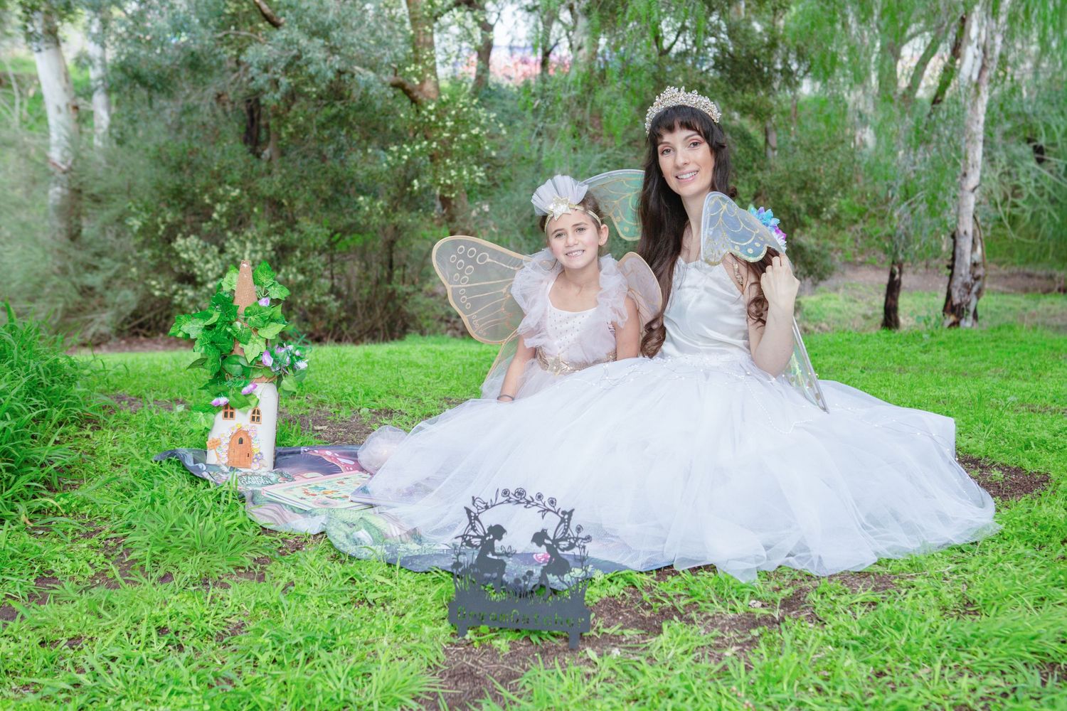 A woman and young girl in white fairy costumes with wings sit together on grass in a park.