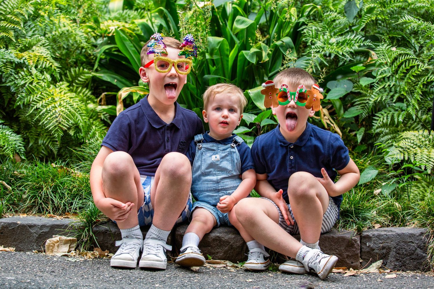 Three boys wearing novelty sunglasses make funny faces while sitting together in a garden.