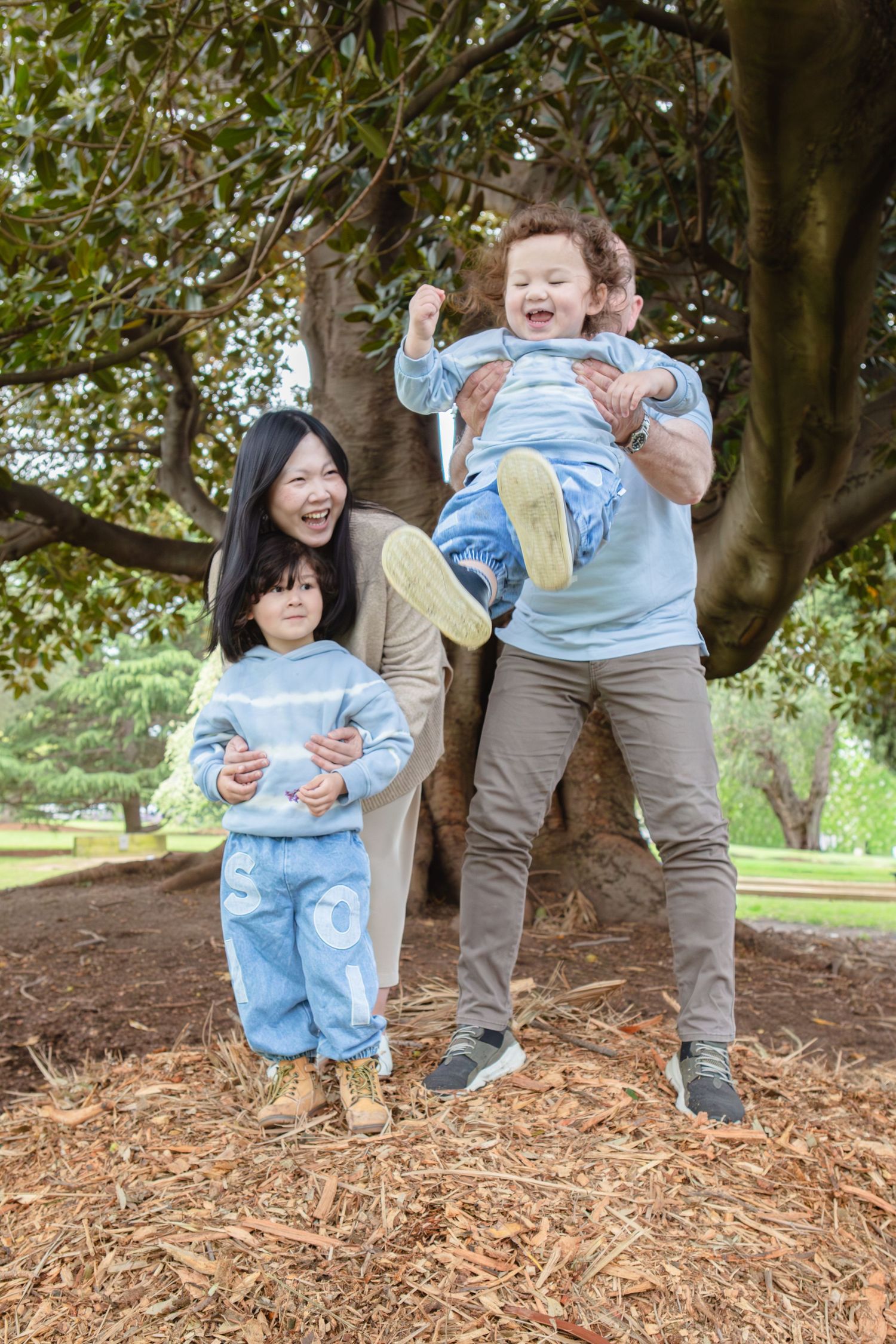 Family enjoying outdoor park time, woman and man lifting laughing child near large tree on wood chip ground.
