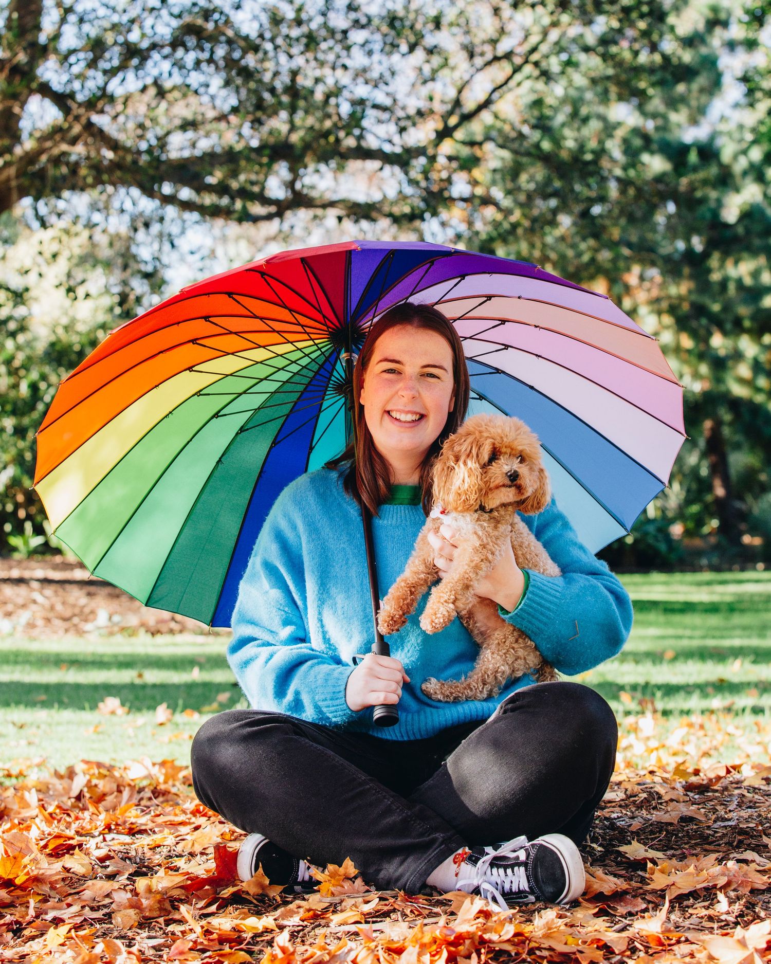 Woman sitting cross-legged on autumn leaves holding a fluffy dog under a colorful rainbow umbrella in a park.