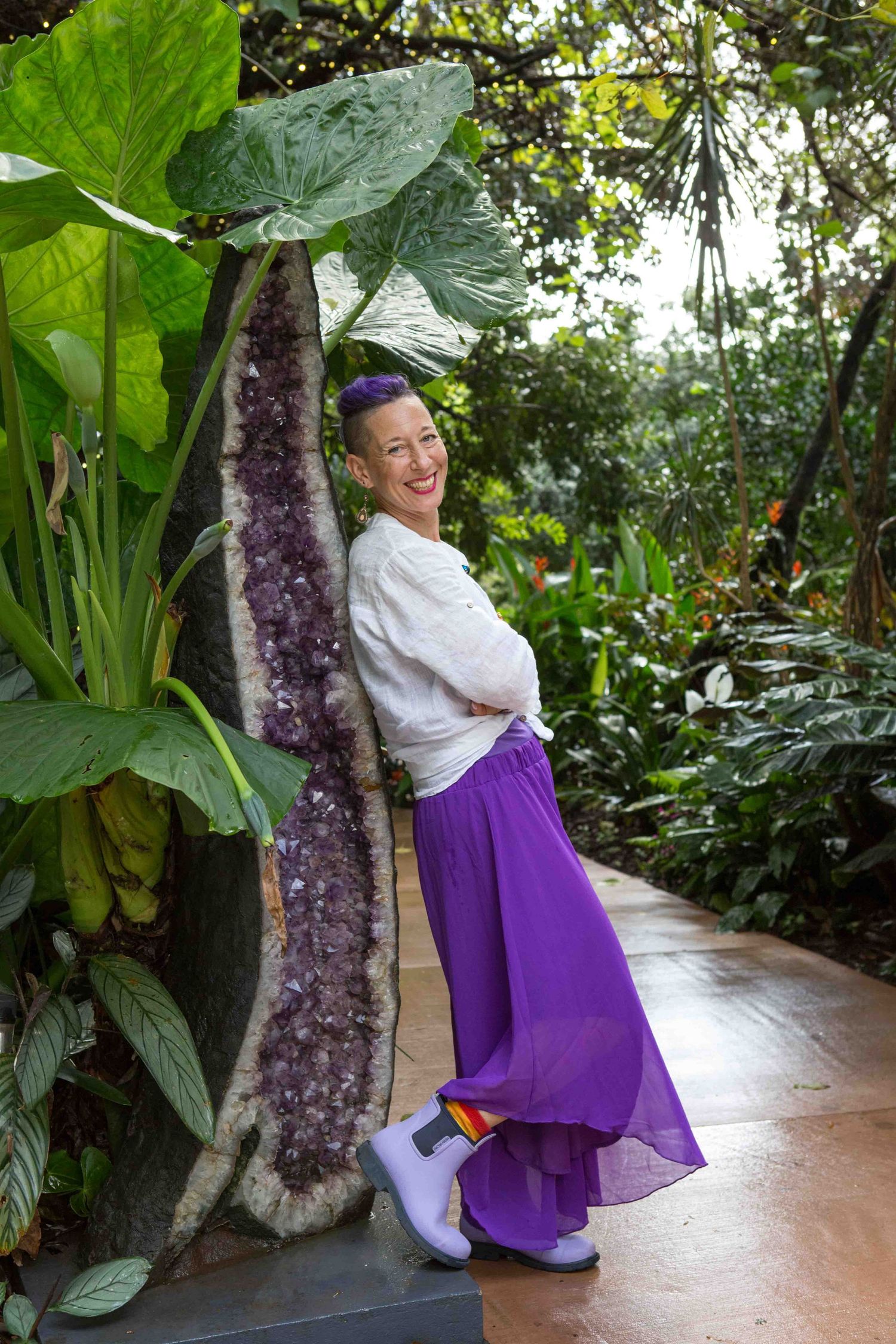 Woman in purple skirt leans against tall amethyst crystal column surrounded by lush tropical garden greenery.