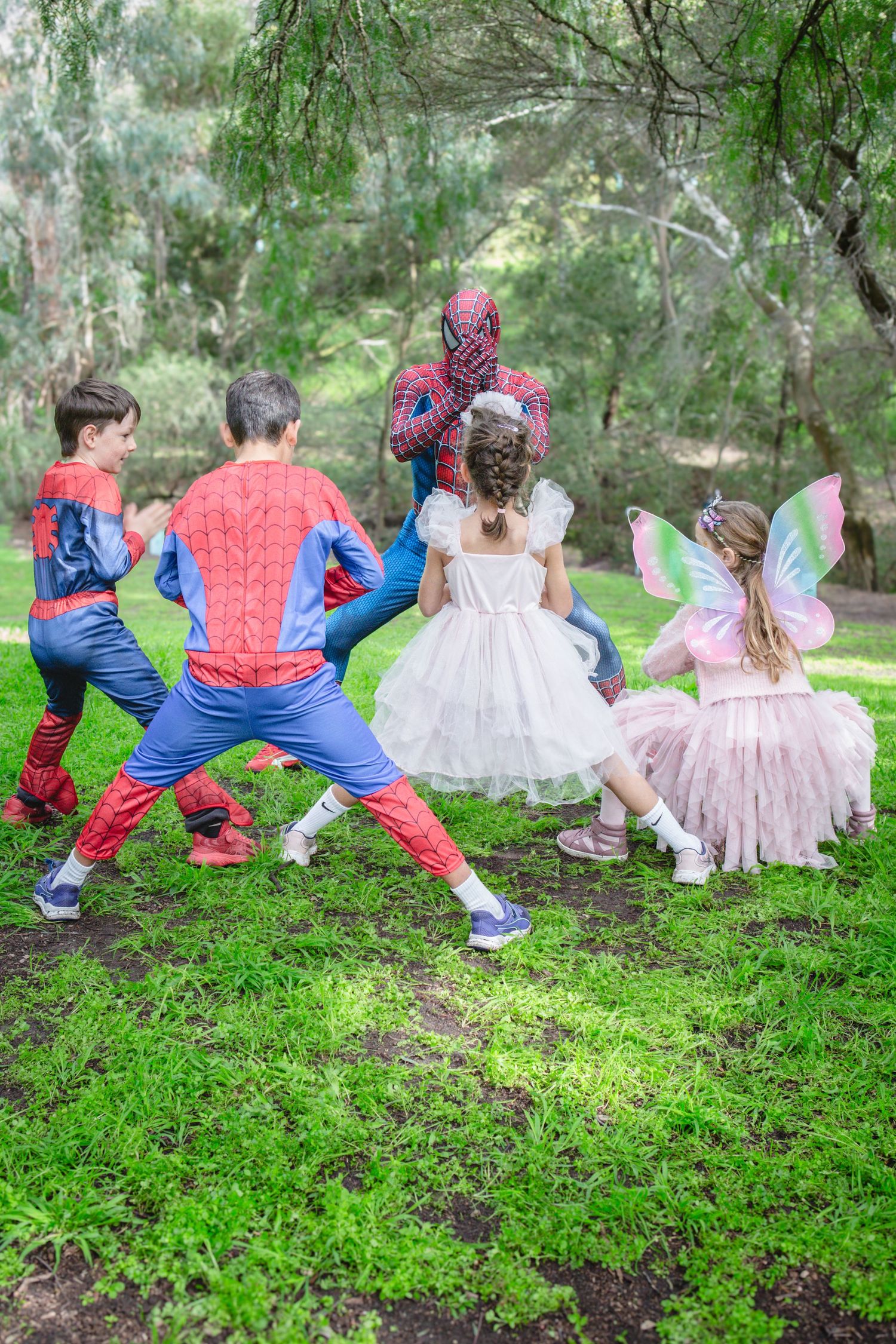 Children in Spiderman and fairy costumes playing together outdoors in a lush green park setting.
