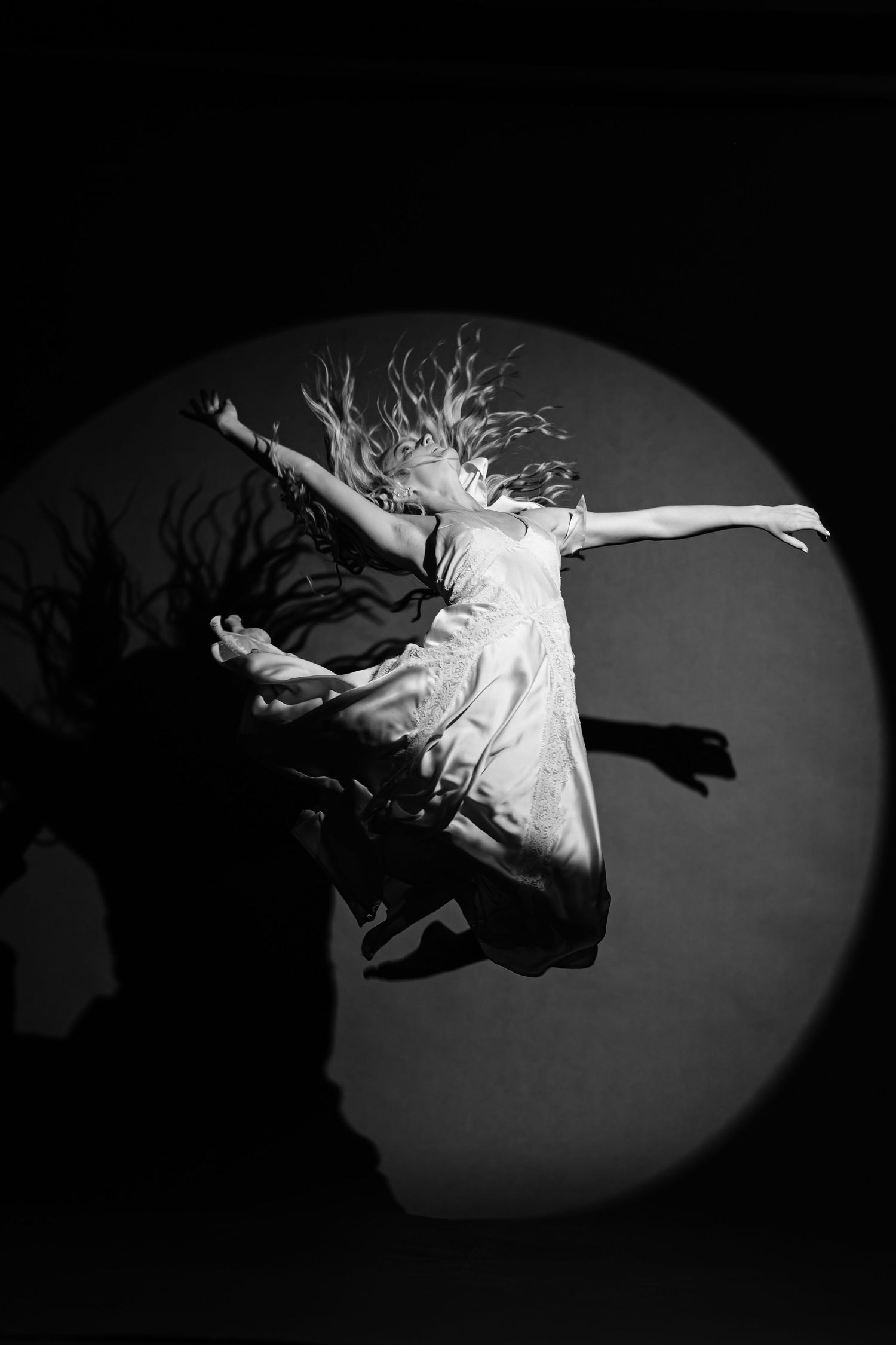 Black and white photo of a dancer in a flowing dress leaping mid-air with arms outstretched inside a circular spotlight.