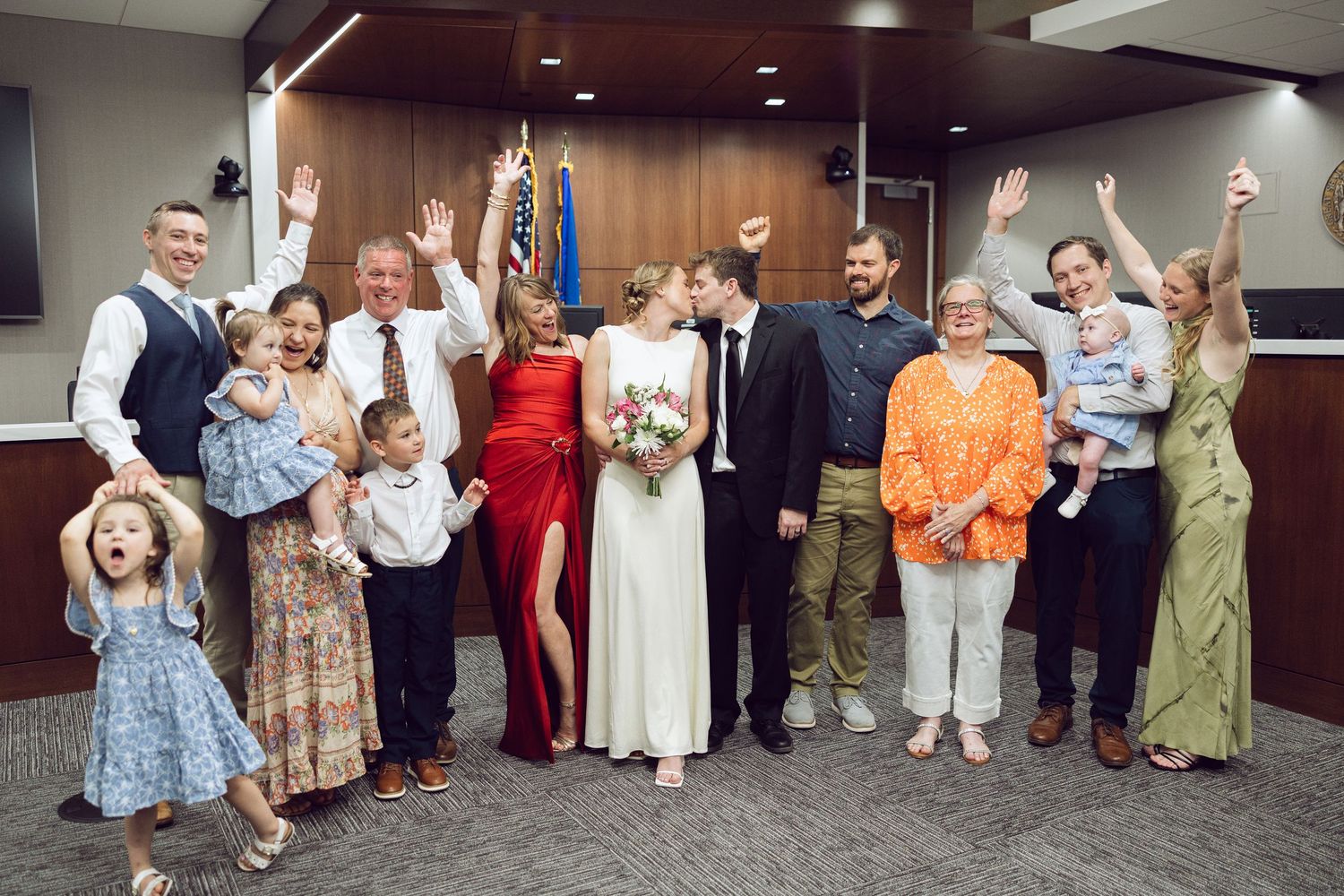 A bride and groom celebrate with joyful family and children, arms raised, in a wood-paneled room after their wedding ceremony.