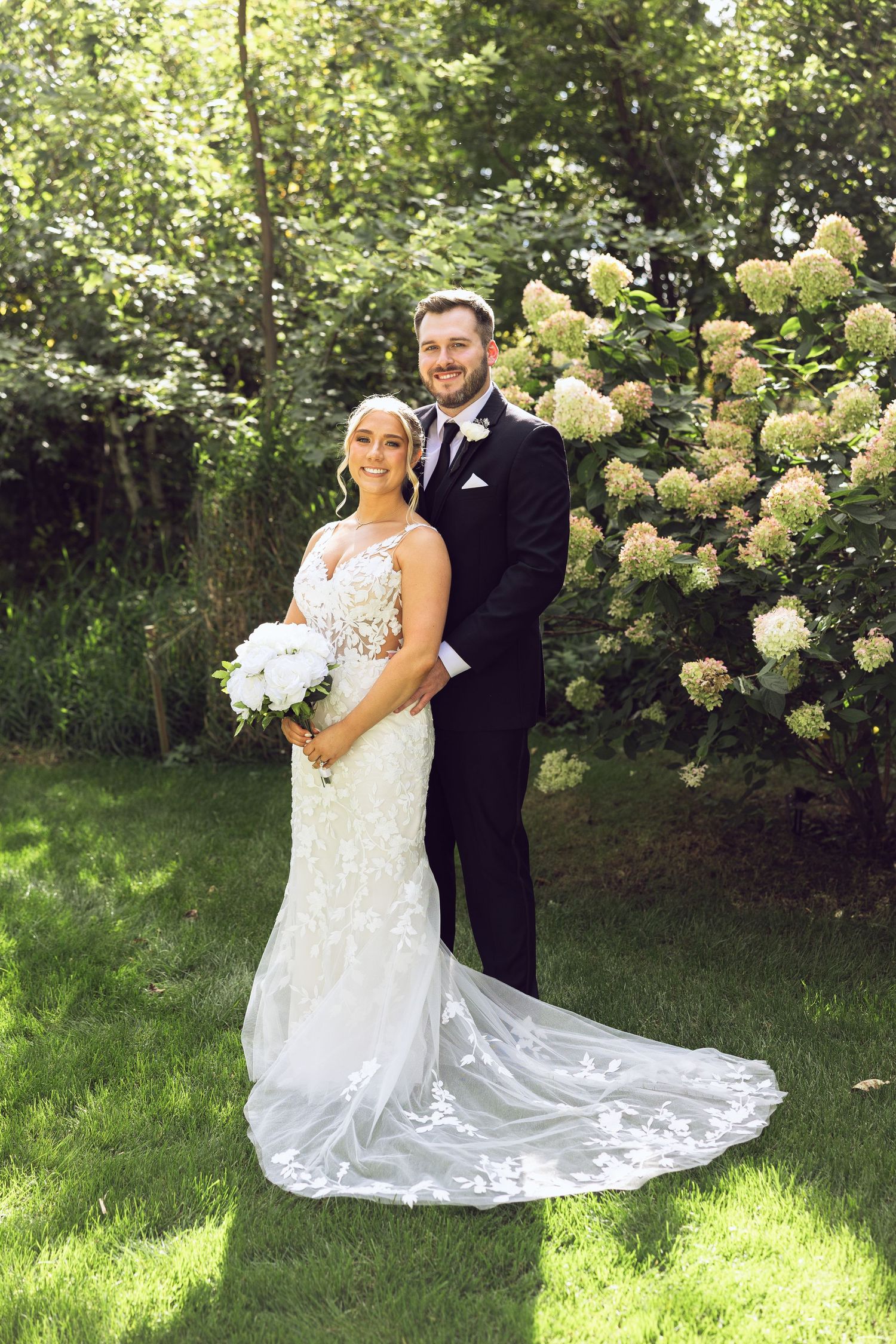 Bride in a lace gown and groom in a black suit pose together outdoors beside blooming hydrangeas on a sunlit lawn.