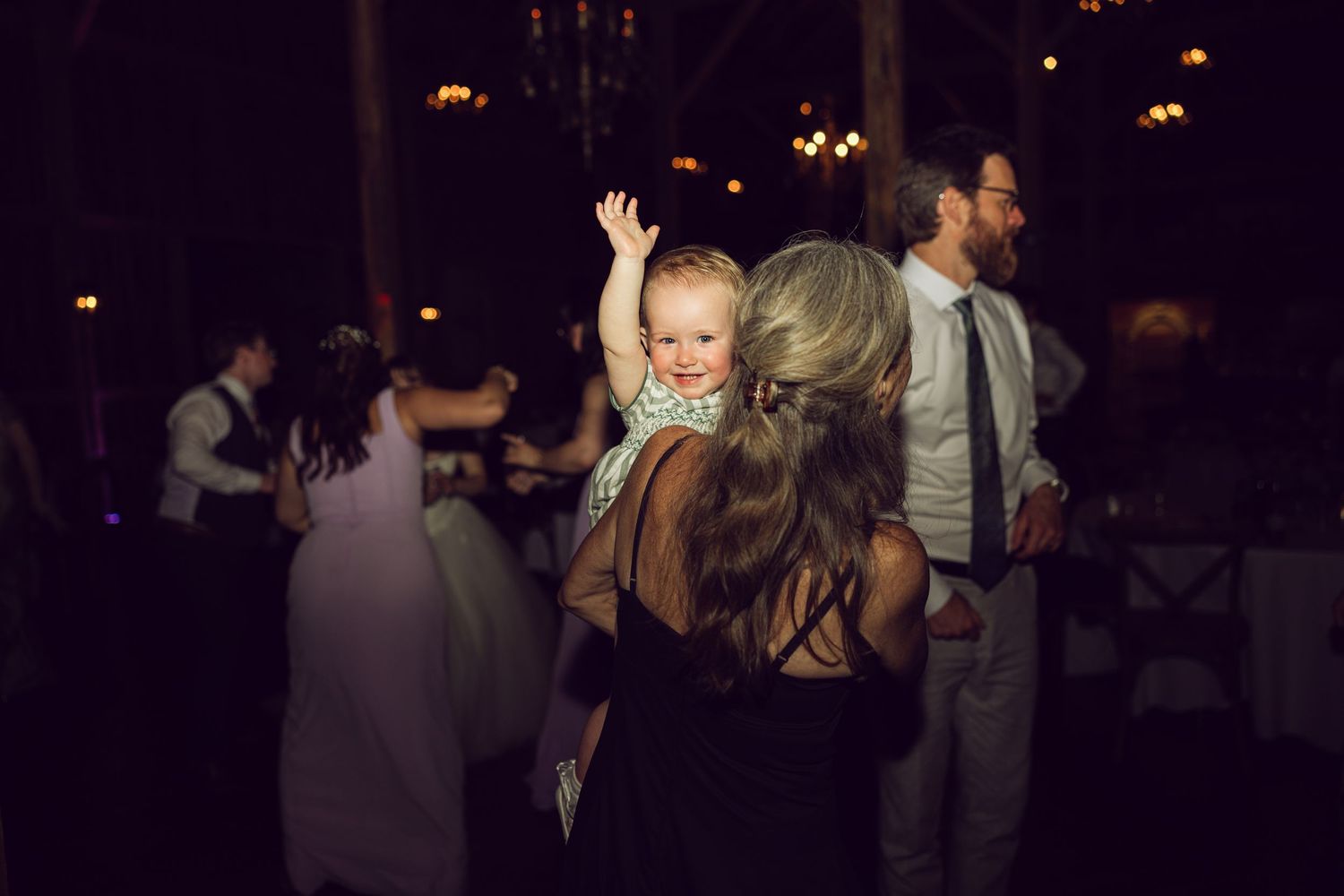 Baby with raised arms looks over a woman's shoulder while guests mingle in a dimly lit wedding reception.